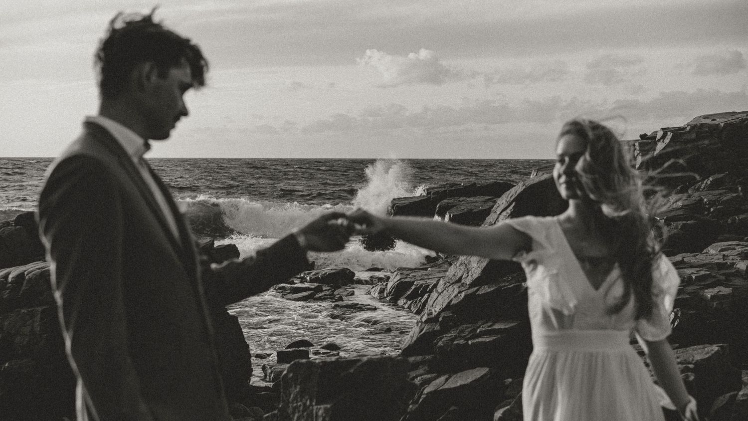 Two people stand on rocky ocean cliffs in wedding attire as waves crash against the shore in black and white.