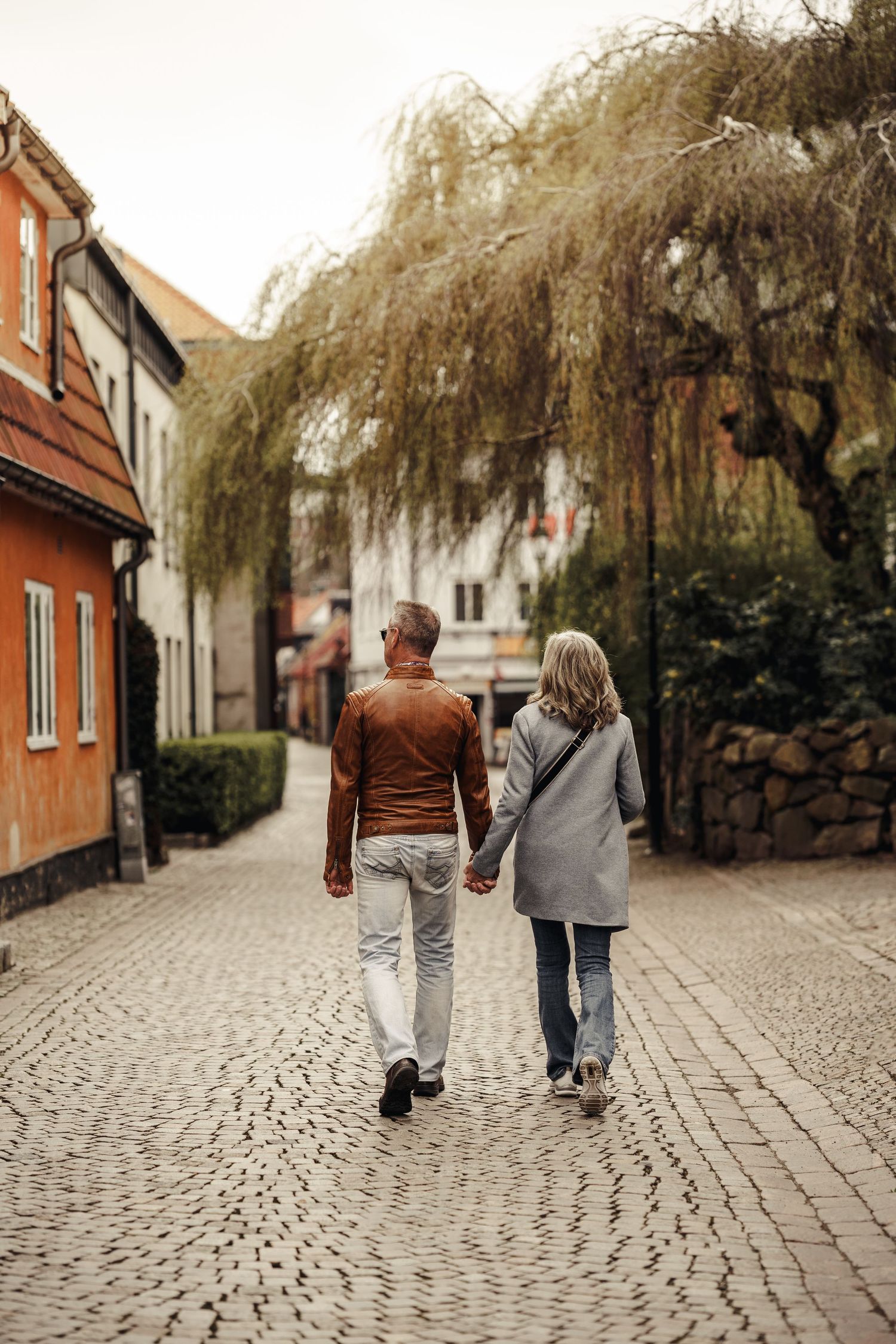 Back view of a couple walking hand in hand down a cobblestone street with a weeping willow tree overhead.