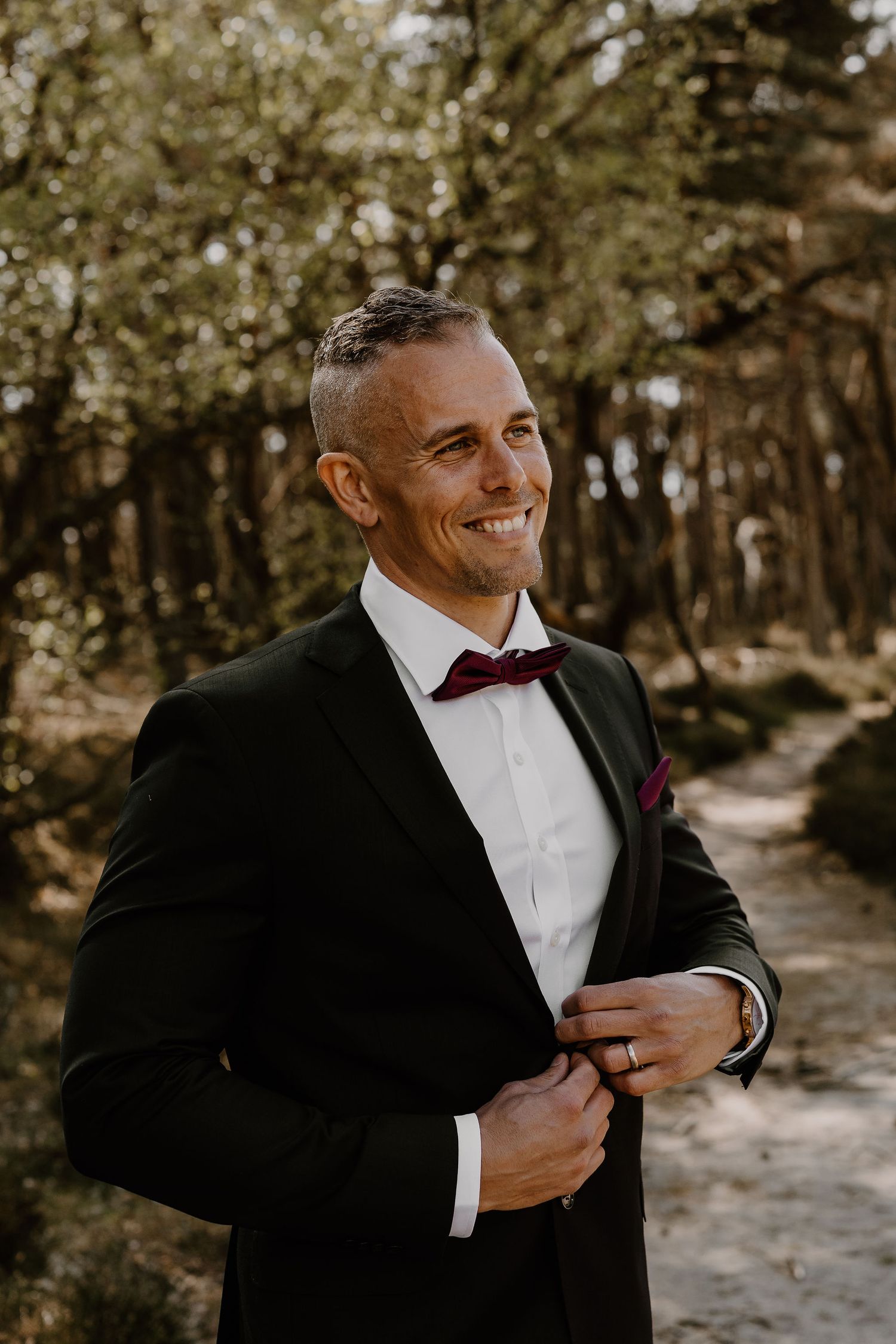A groom in a classic black tuxedo and burgundy bow tie smiles while adjusting his jacket in a wooded setting.