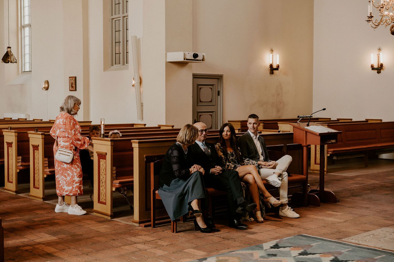 People sit together on wooden pews inside a historic church with warm lighting and white walls.