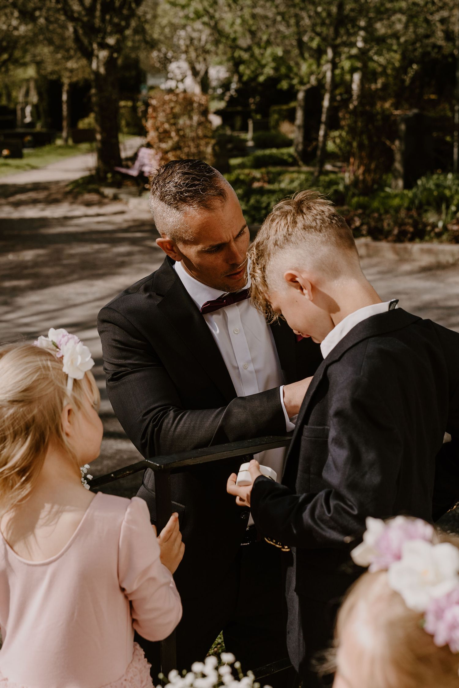 A touching moment before the wedding ceremony in a sunlit outdoor garden setting with formal attire and floral details.