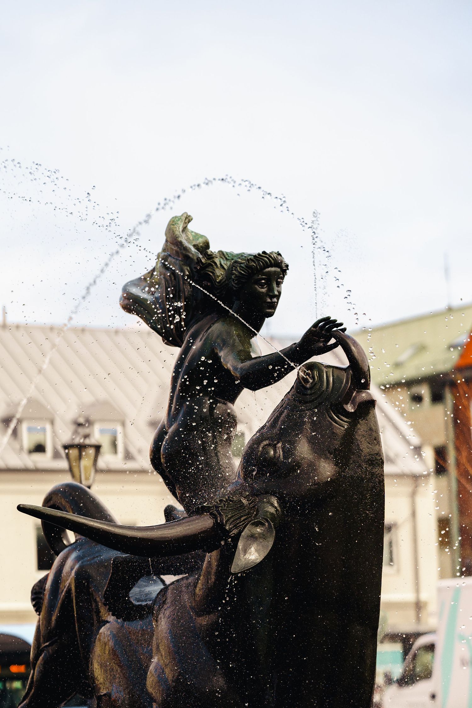 Silhouette of a bronze fountain sculpture with water spraying dramatically in sunlight against urban architecture.