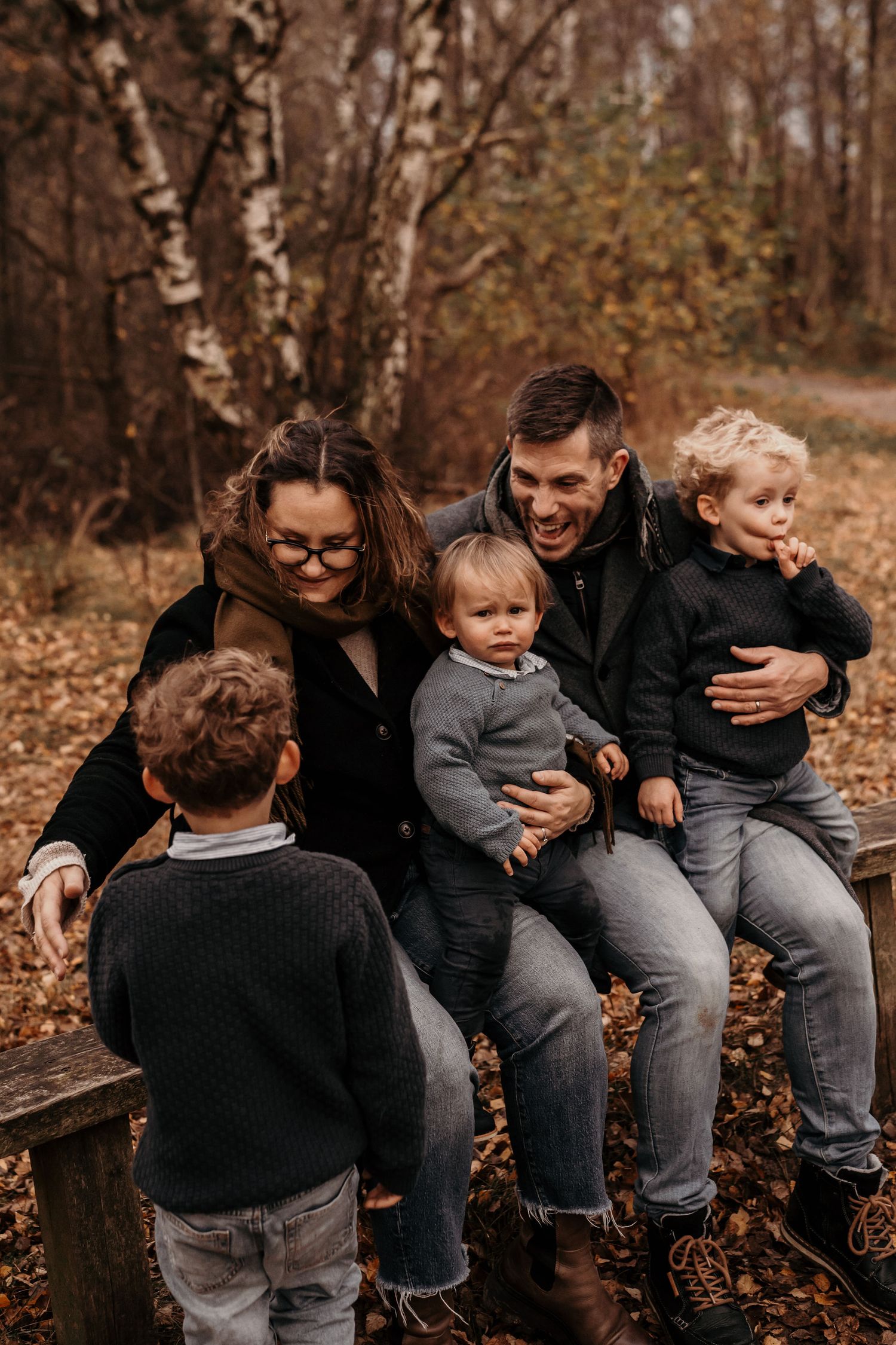 A family shares a joyful moment sitting together on a wooden bench surrounded by autumn leaves in the woods.