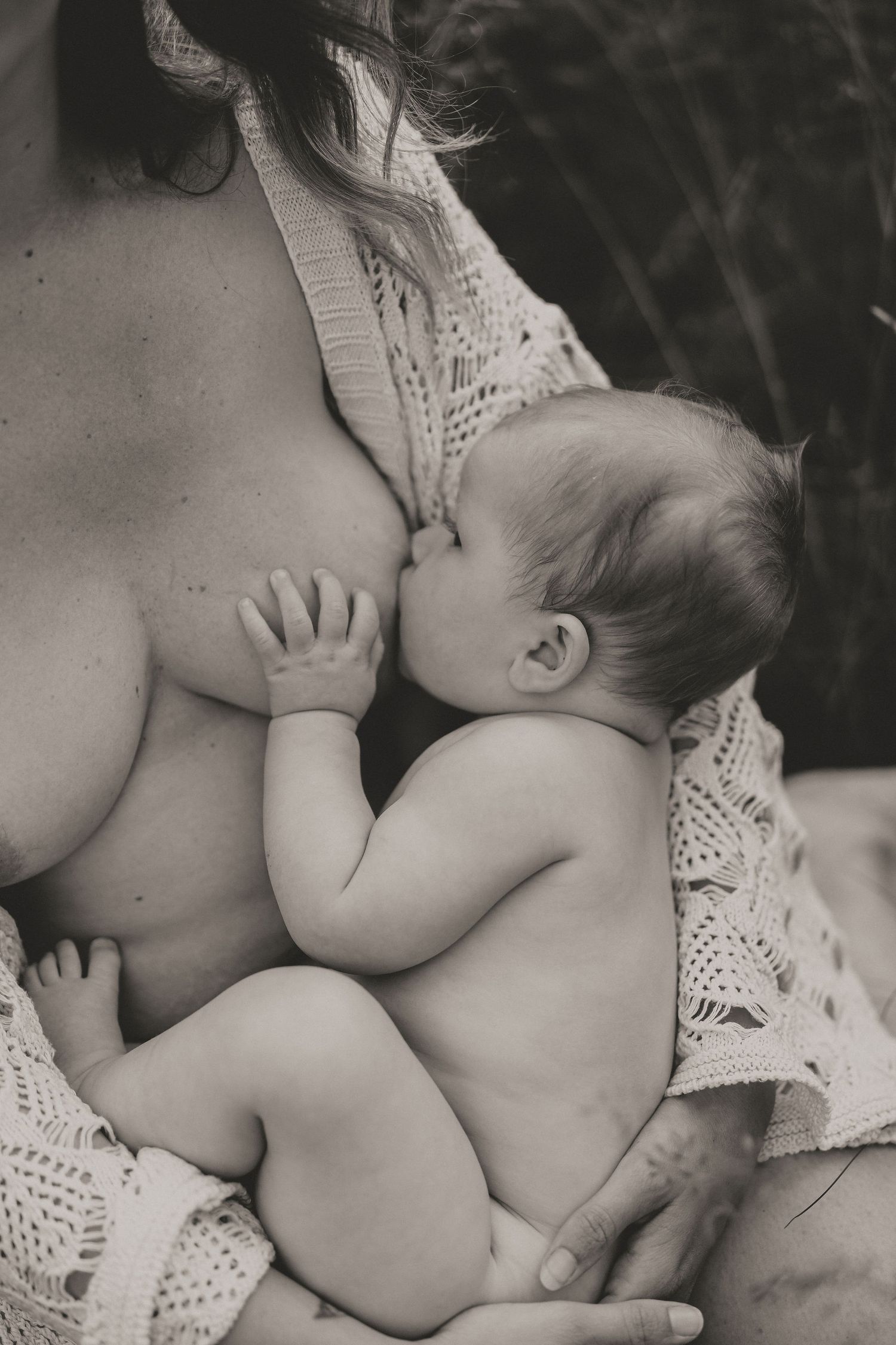 Black and white intimate photograph of a baby breastfeeding, draped in delicate lace fabric.