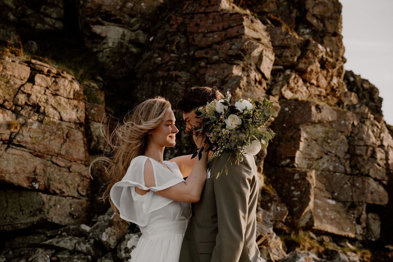 A couple embraces during their wedding photoshoot against dramatic rocky cliffs while holding a romantic bouquet of flowers.