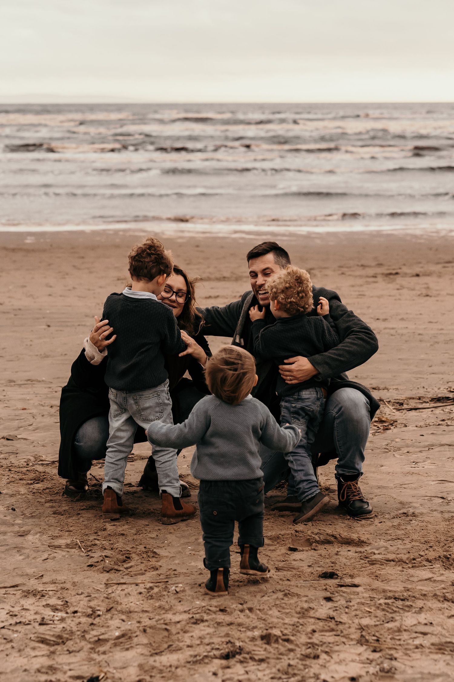 A group huddles together on a cloudy beach while a small child in gray stands in front of them.