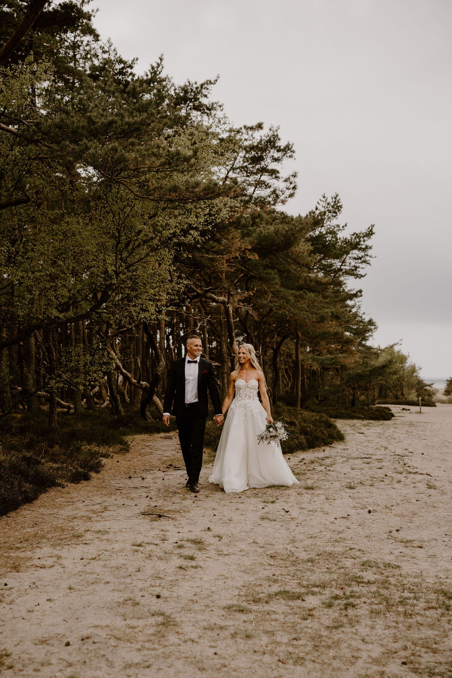 A romantic couple walk together on a sandy beach path lined with tall pine trees on an overcast day.