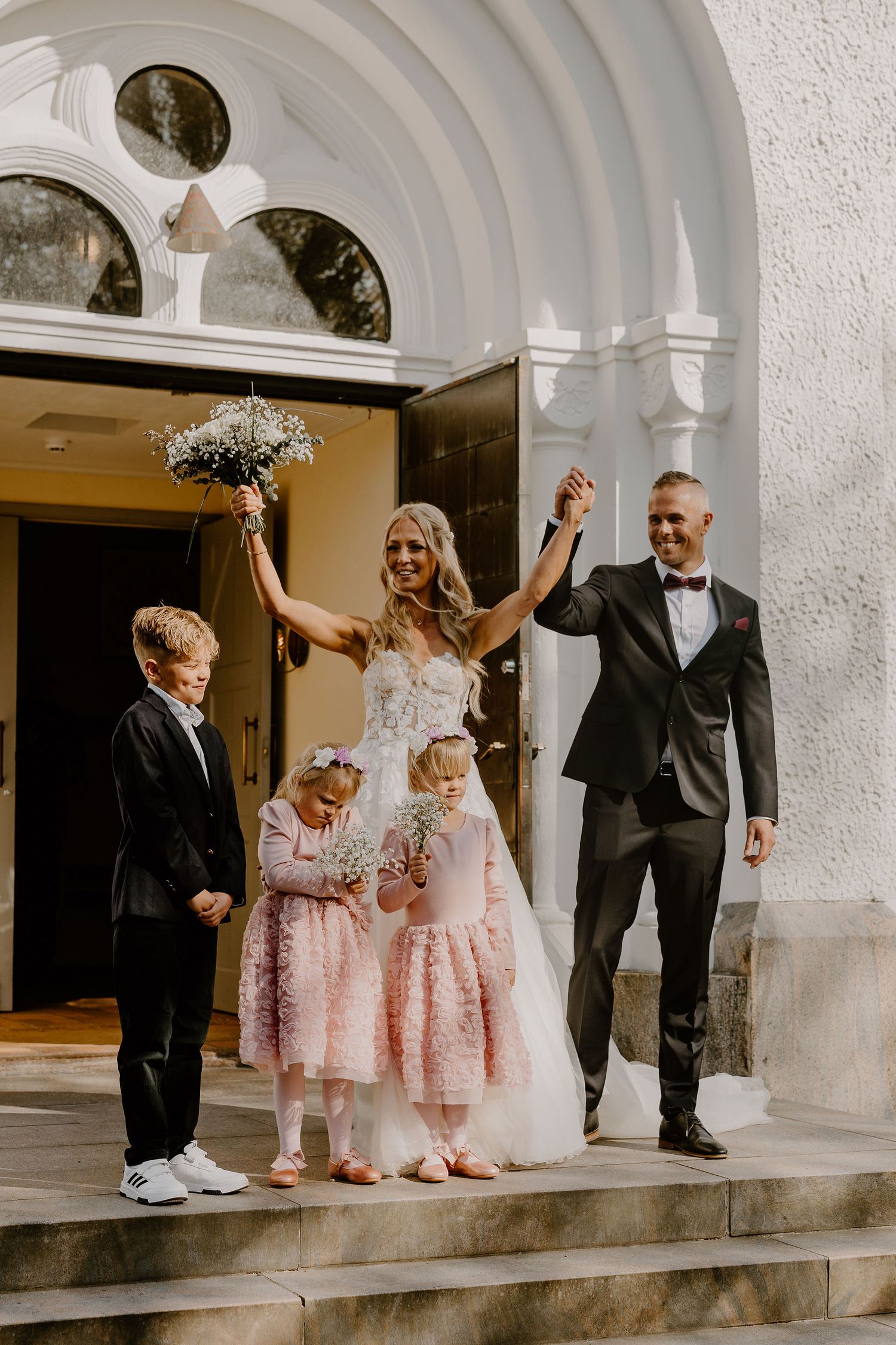 Newlyweds celebrate with raised arms on church steps alongside young attendants in pink dresses and formal attire.