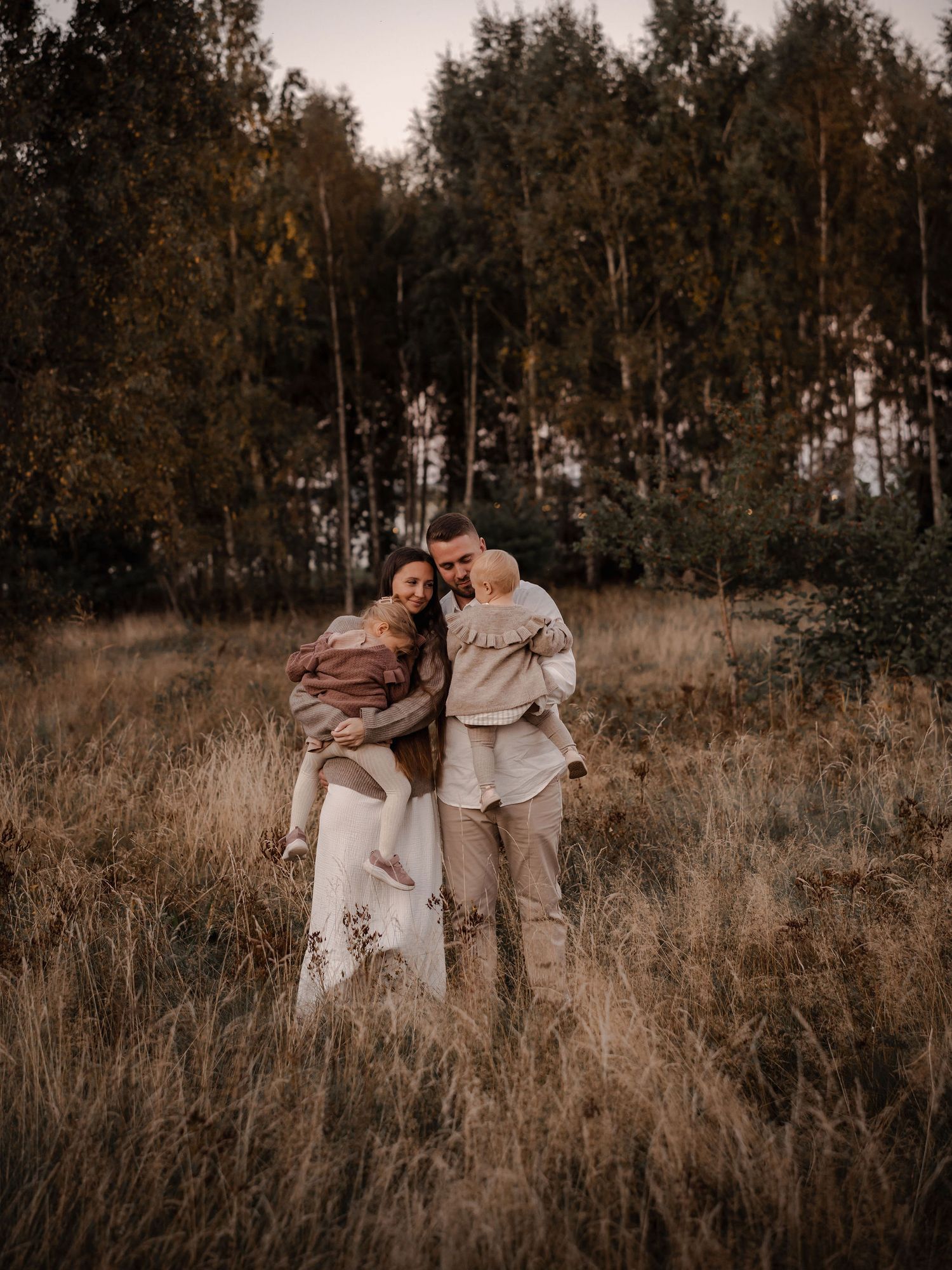 A loving family stands together in a golden field with tall trees silhouetted behind them.