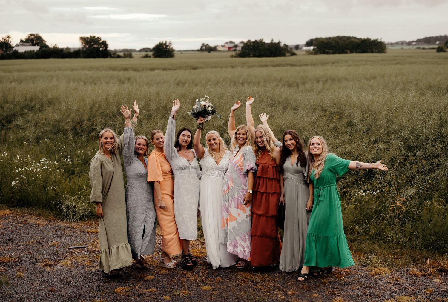 A group in assorted flowing maxi dresses pose joyfully with raised arms on a dirt path amid rustic countryside fields.