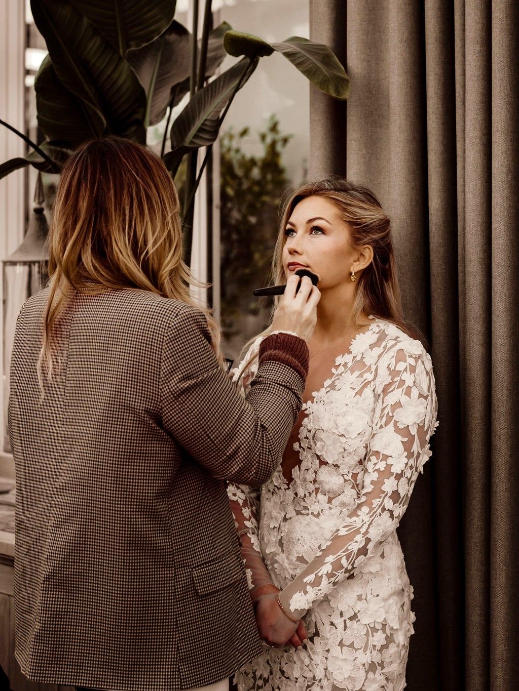 A makeup artist applies finishing touches to a bride wearing an elegant white lace long-sleeved wedding dress.