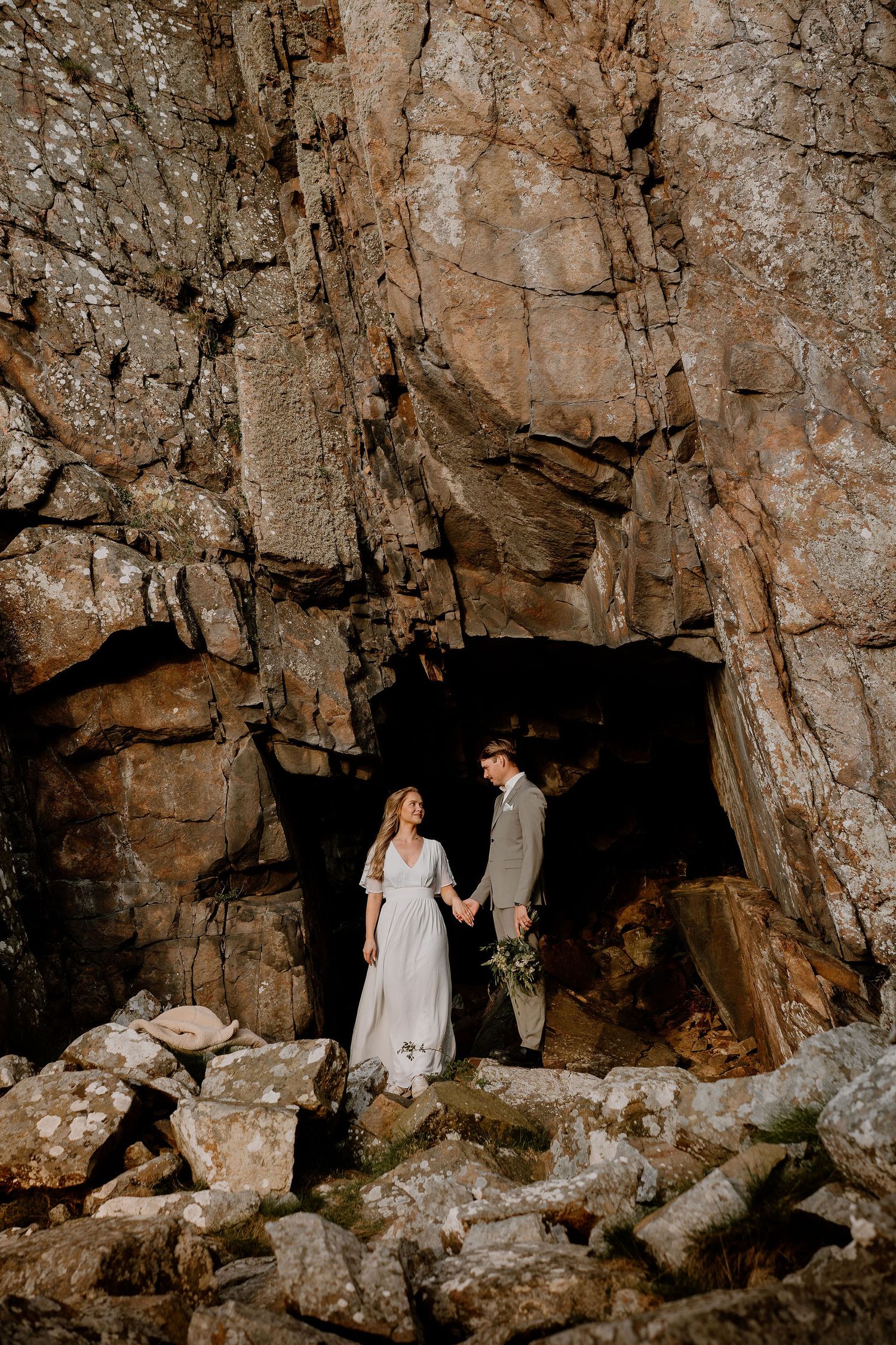 Two people in wedding attire stand at entrance of dramatic mountain cave surrounded by rugged rock formations.