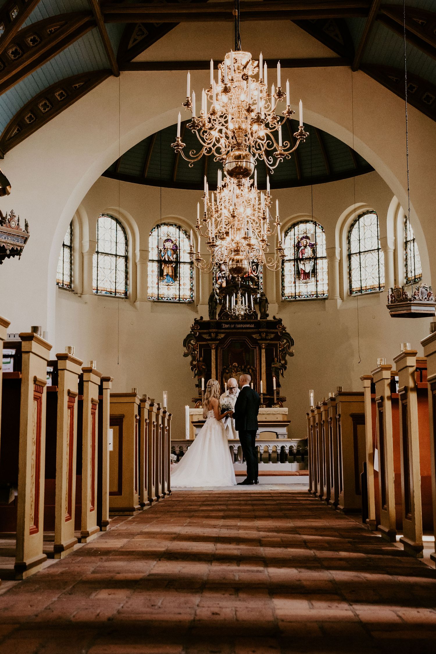 Elegant church interior with grand crystal chandelier and wooden pews leading to ornate altar under arched windows.