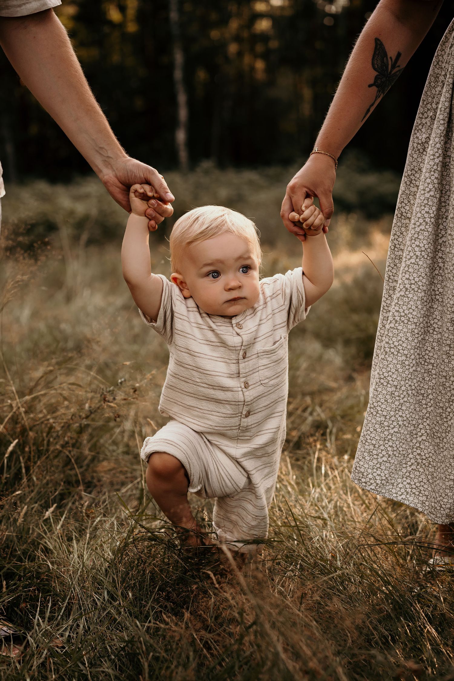 Small child learning to walk while holding parents' hands in a rustic outdoor setting.