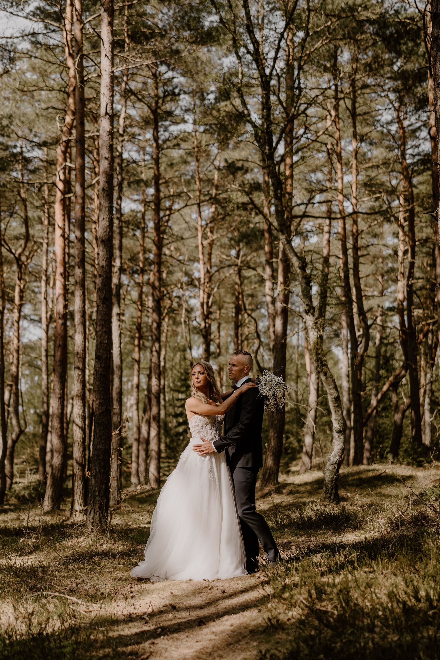 A couple in wedding attire share an intimate moment on a sunlit forest path surrounded by tall pine trees.