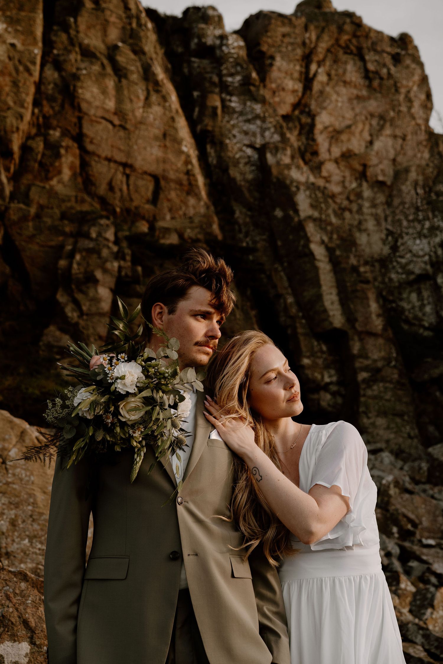 Couple in wedding attire posing against dramatic rocky cliffs at sunset creating moody atmosphere.