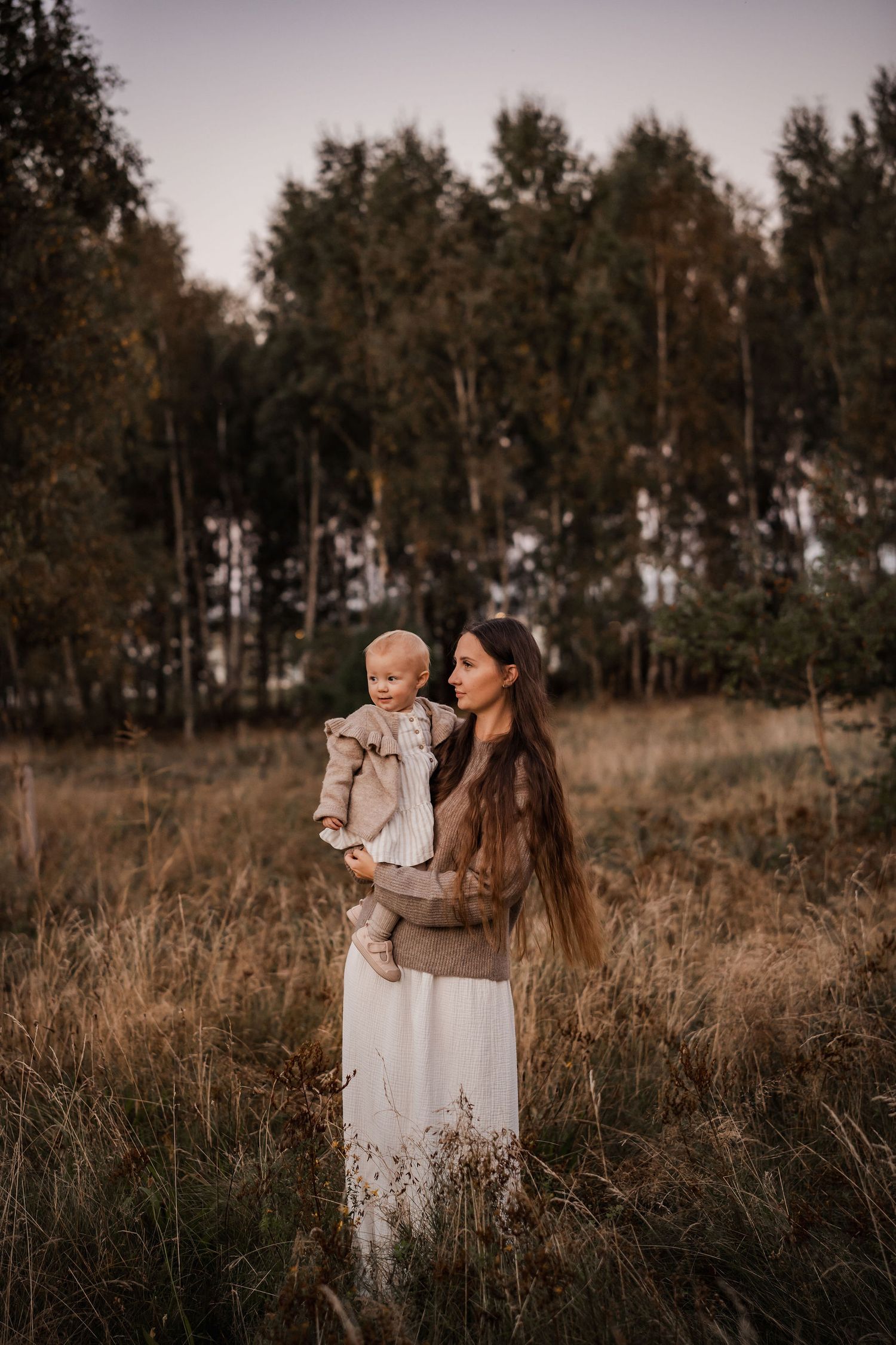 Woman in white dress holds small child while standing in field at sunset with forest backdrop.