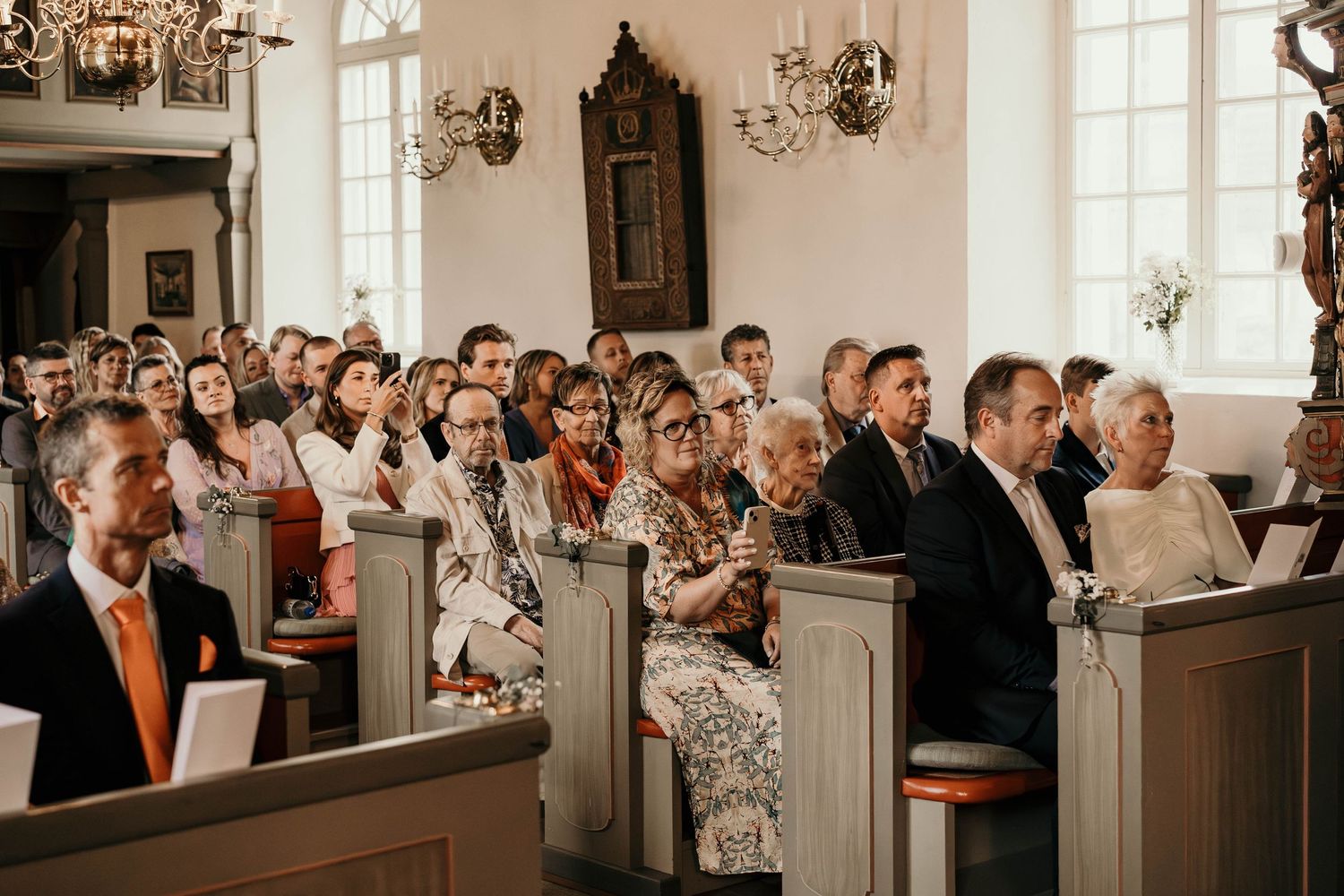 Wedding guests sitting in traditional wooden pews of a bright church with elegant chandeliers and wall sconces.