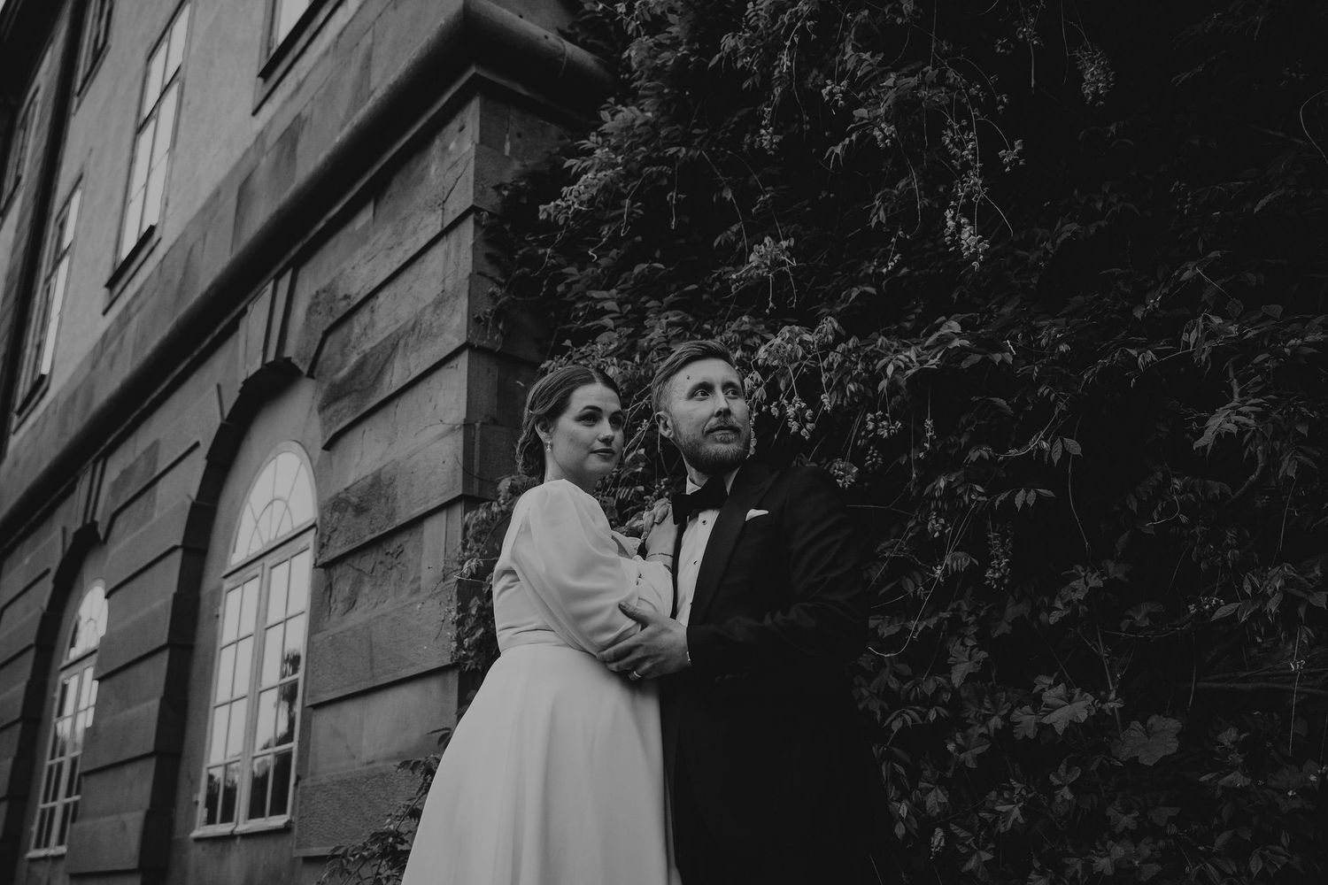 A black and white photo shows a couple cuddling outdoors against a textured wall exterior setting.