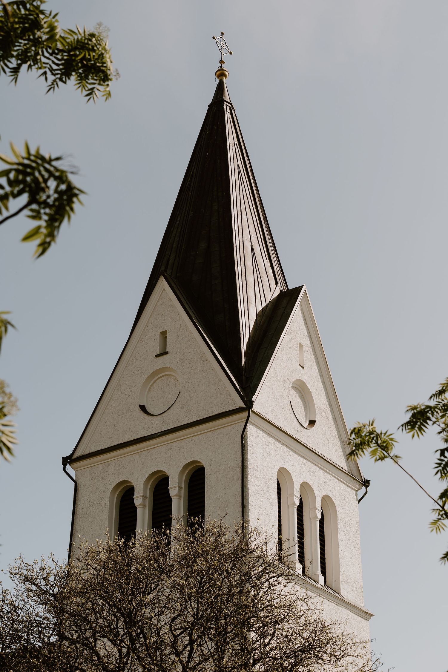 Tall white church steeple with pointed spire and arched windows rises against cloudy sky, framed by tree branches.