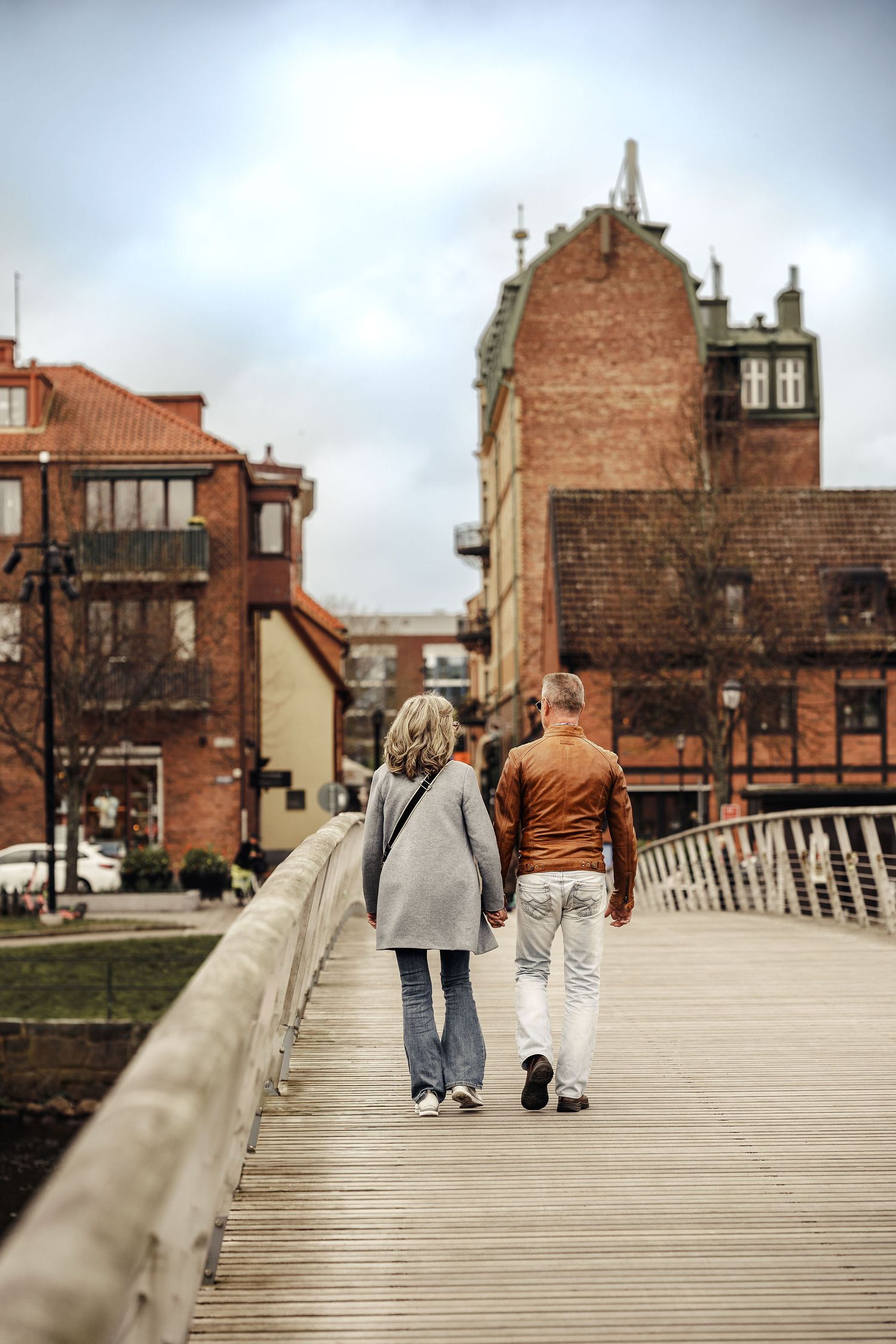Two figures walk together on a wooden boardwalk in an urban setting with brick buildings and cloudy skies in the background.