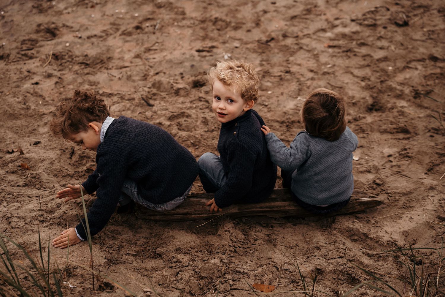 Three children sit together on a muddy slope in casual clothing during an outdoor adventure.