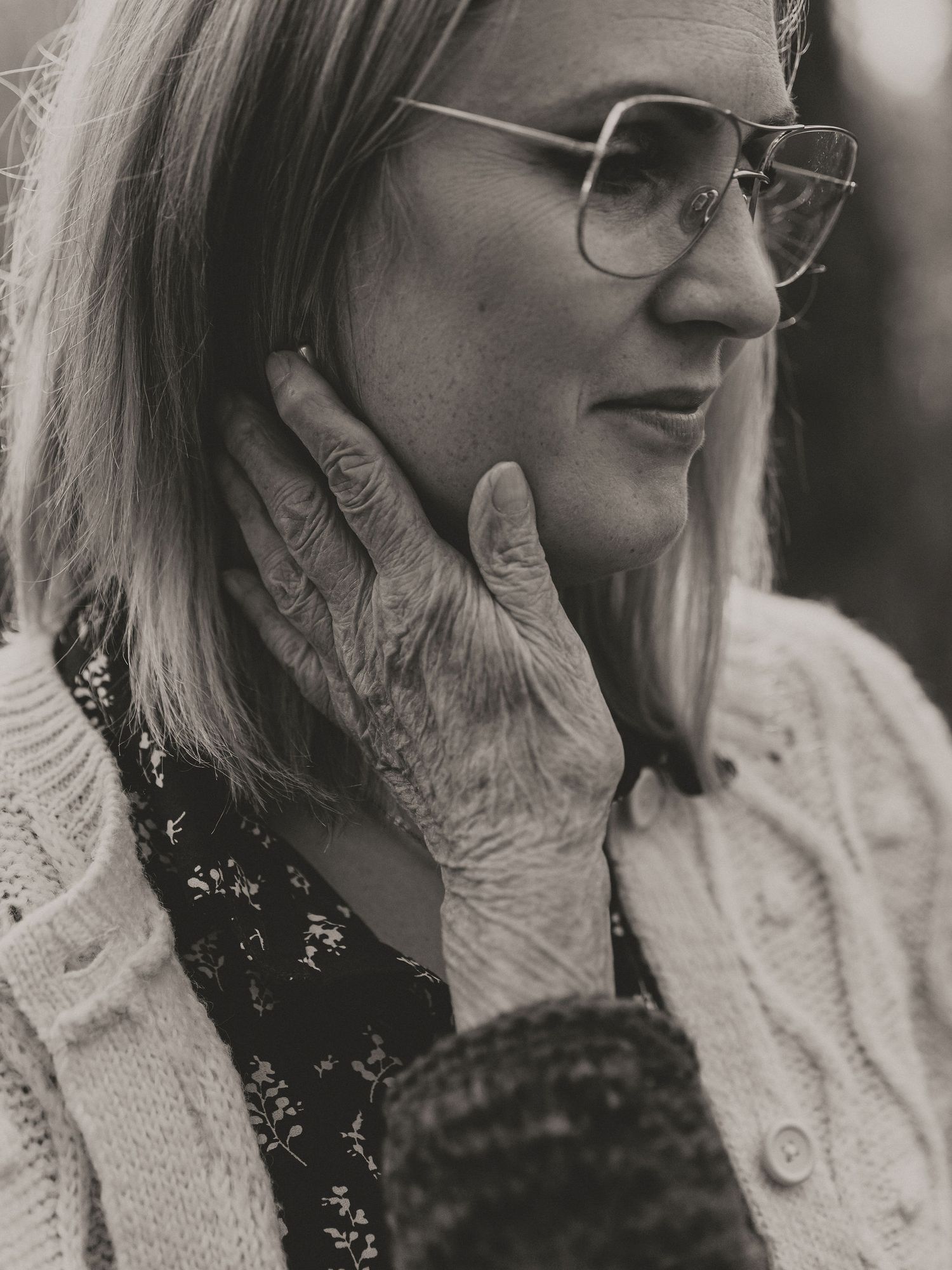 Black and white close-up photo showing an elderly hand gently touching a younger person wearing glasses and a cardigan.