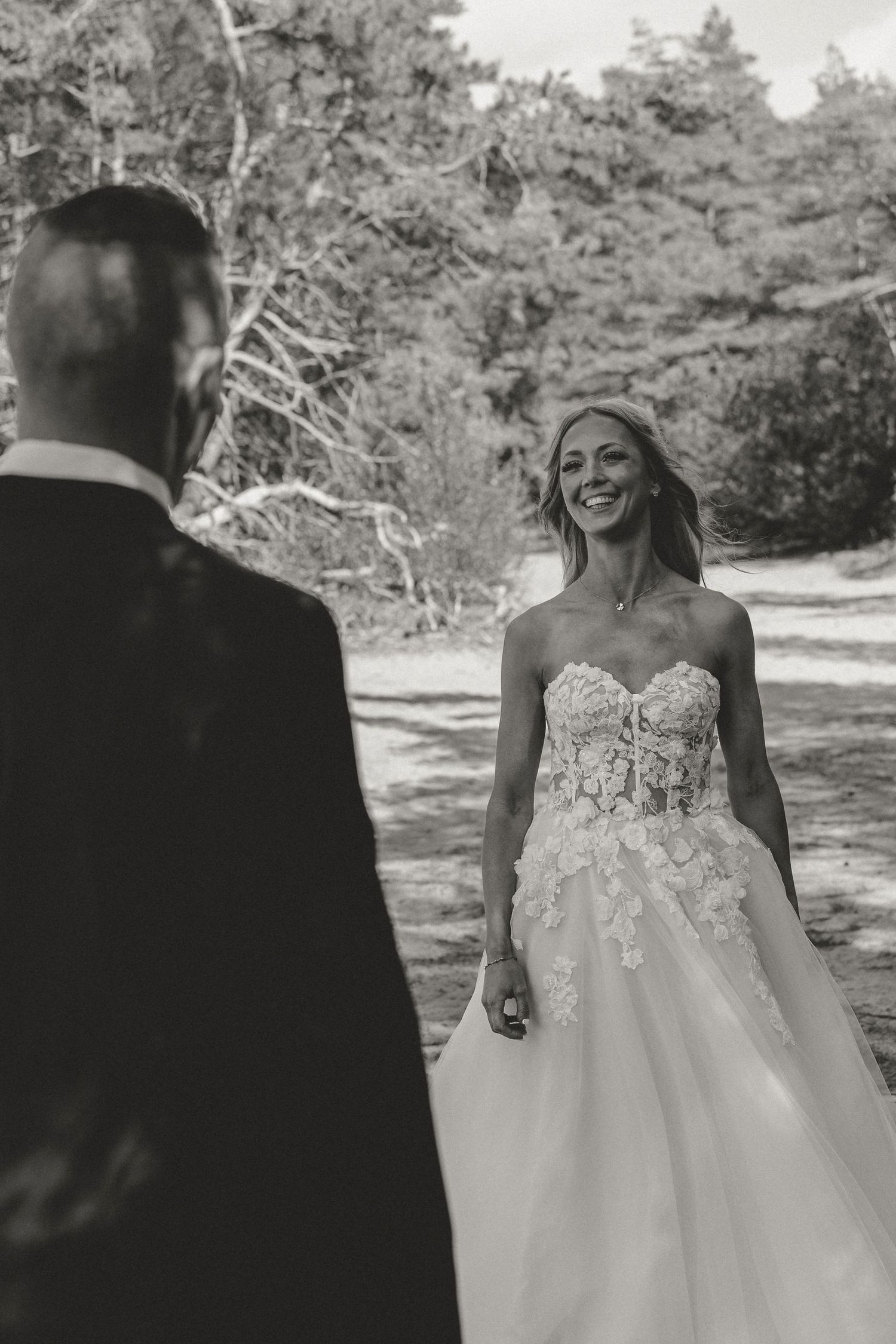 Black and white photo of a bride in a lace strapless wedding gown sharing a candid moment outdoors with wedding trees behind.