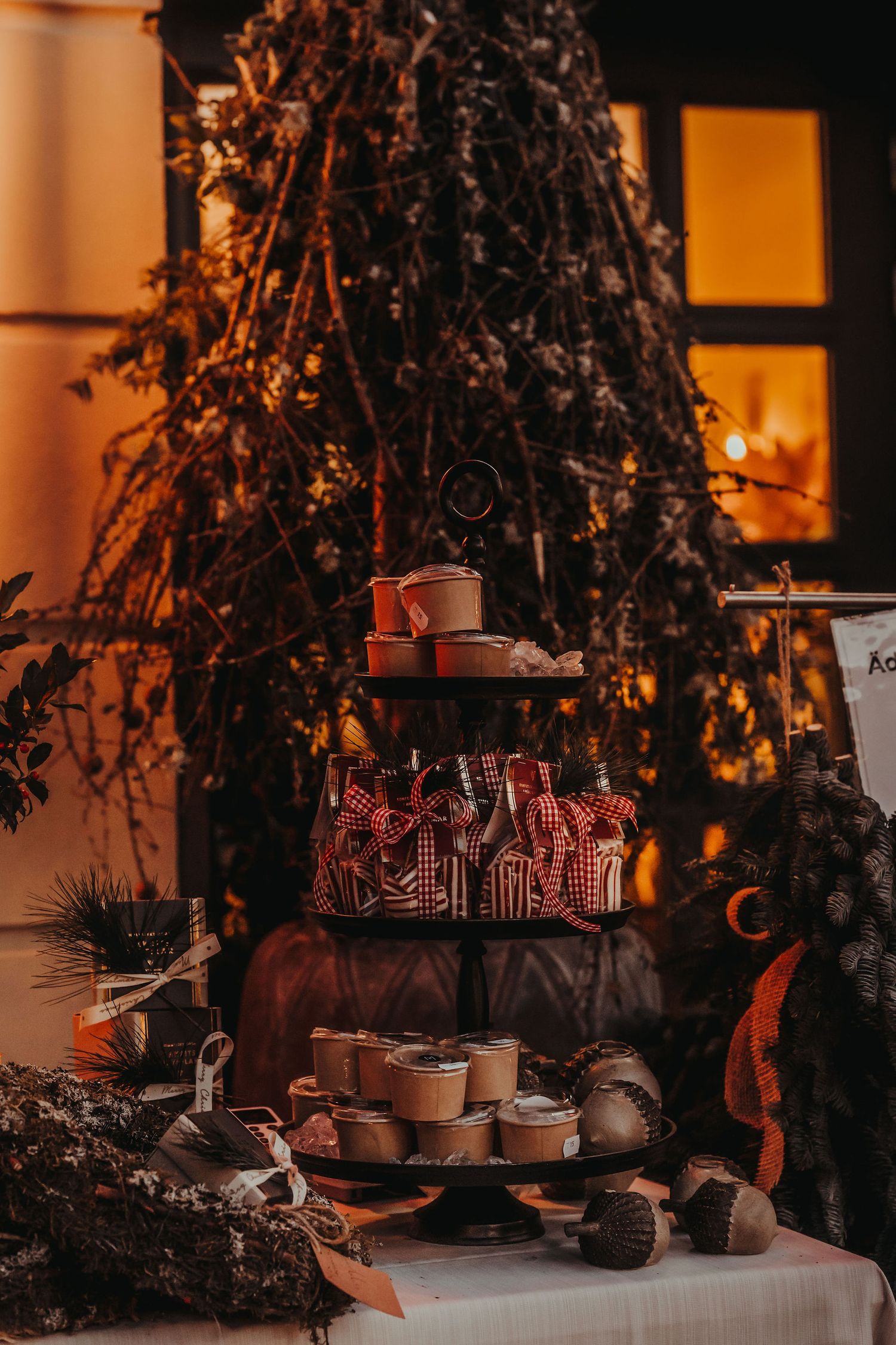 Cozy Christmas dessert display with tiered stand holding gingerbread houses and treats against warm glowing window setting.
