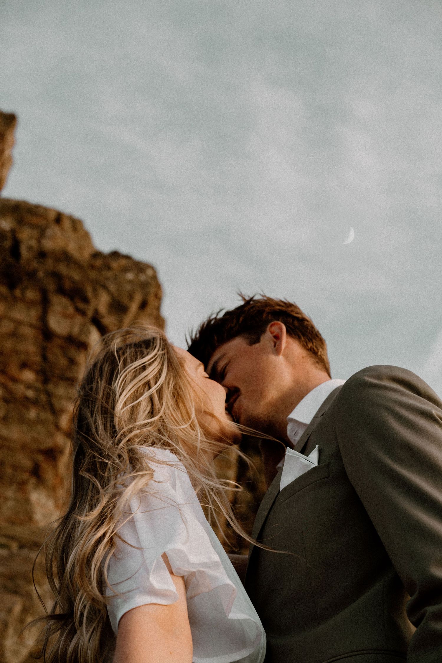 A romantic couple share a kiss against a rugged cliff backdrop with a crescent moon in the overcast sky.