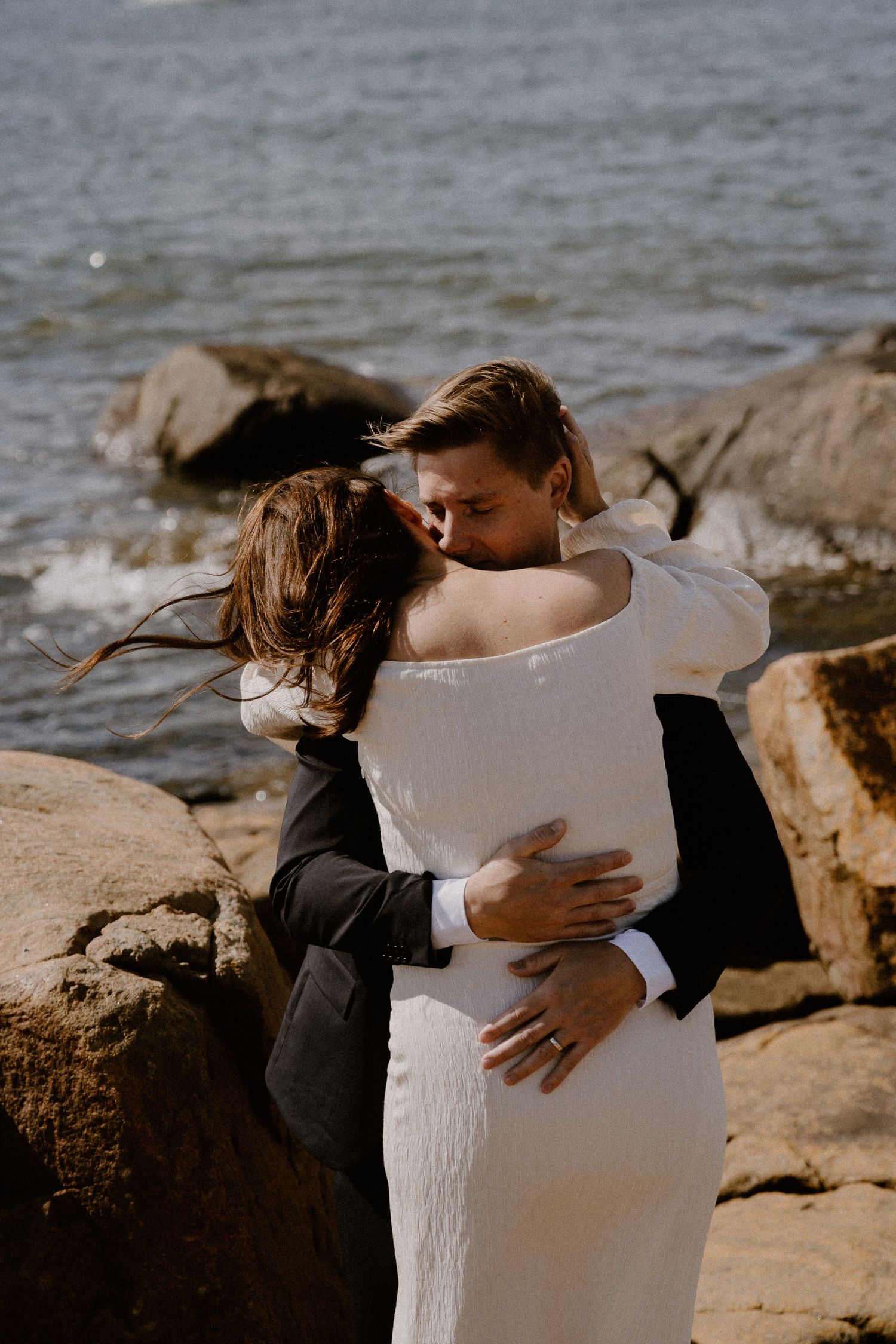Romantic beachside embrace between a couple in formal attire with waves and rocks in the background.