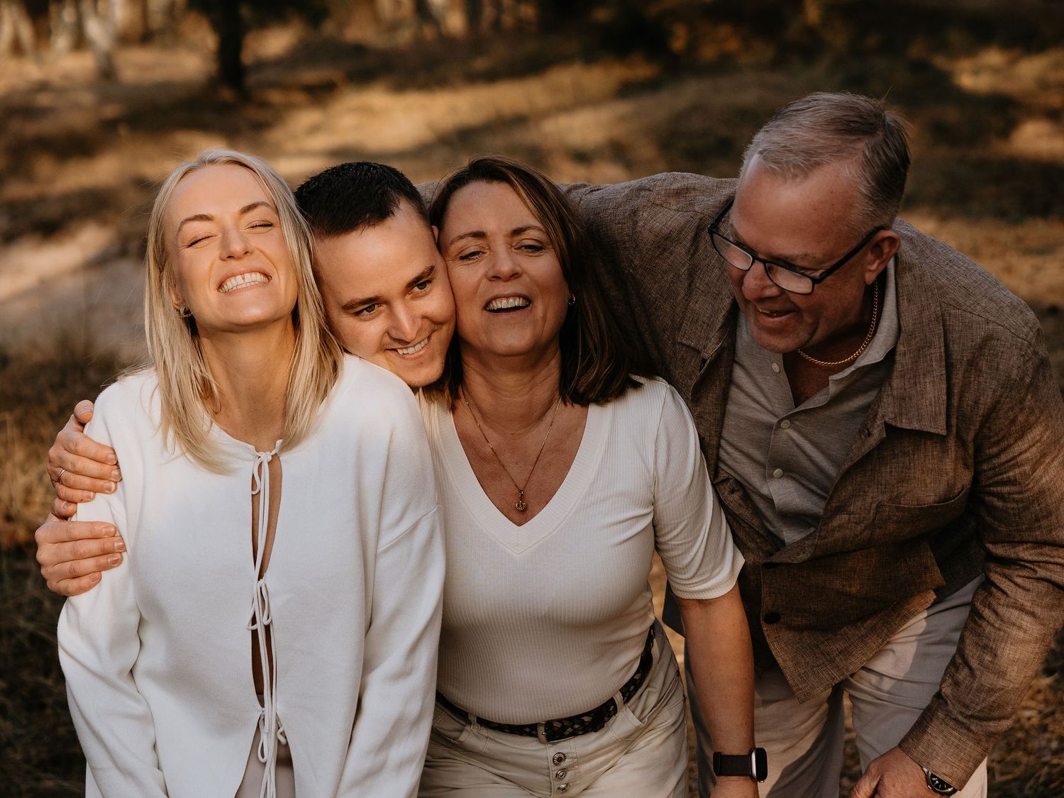 Four people in white tops laughing and embracing together in a warm autumn setting during golden hour.