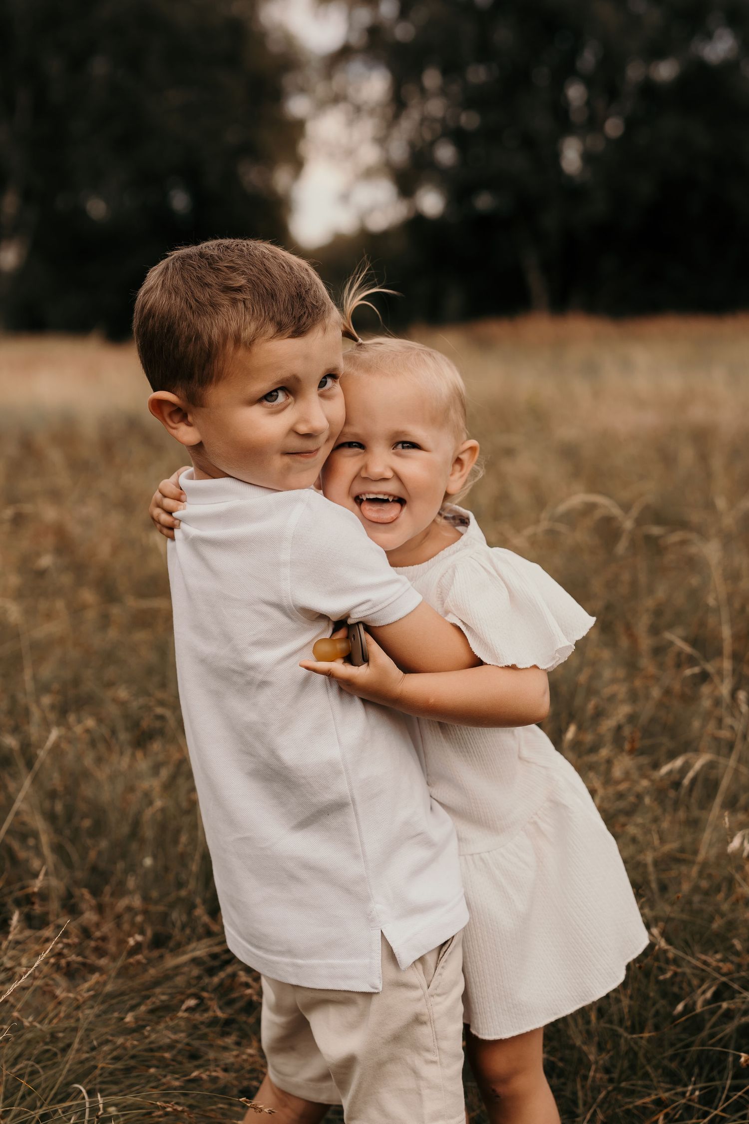 Two children in white outfits sharing joyful hug in golden field.