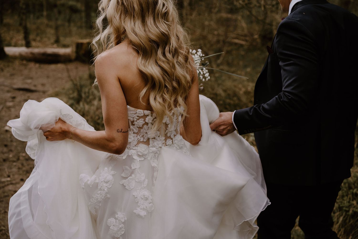 Back view of bride in white floral lace wedding dress holding skirt while walking with groom in dark suit outdoors.