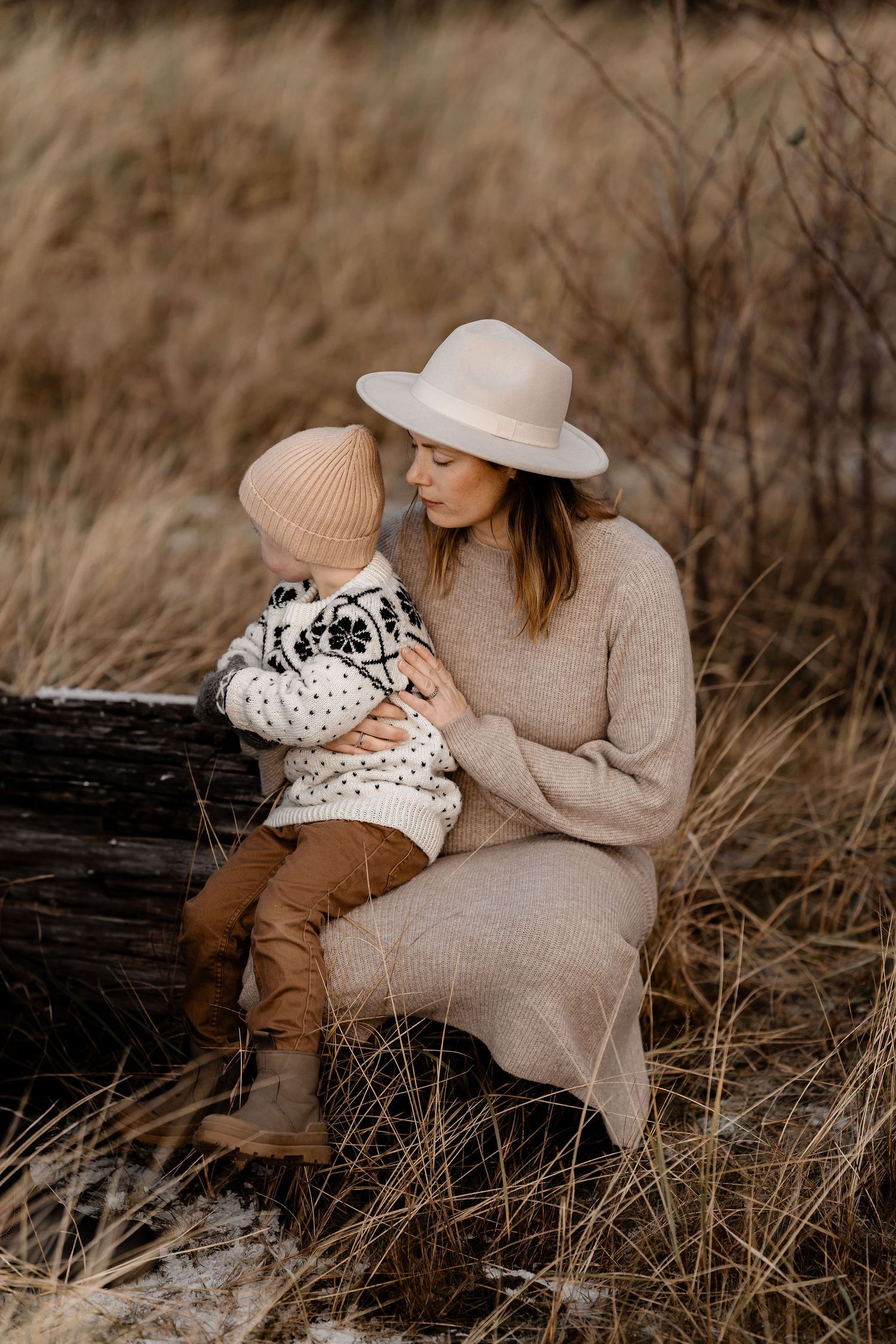 Cozy autumn portrait in earth tones showing tender moment between parent and child on rustic wooden log in tall grass.