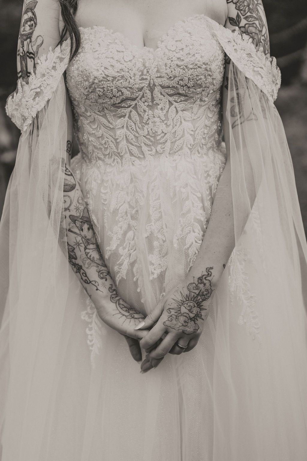 A close-up of an elegant wedding dress with intricate lace details, sheer sleeves and tulle fabric, shown in black and white.