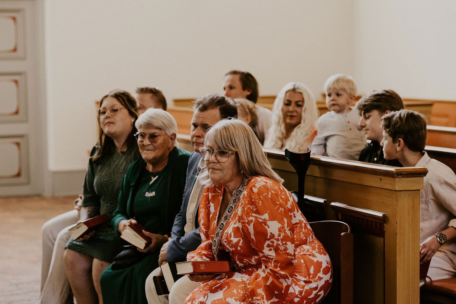A group of people sit attentively in wooden church pews wearing colorful clothing in a bright interior space.