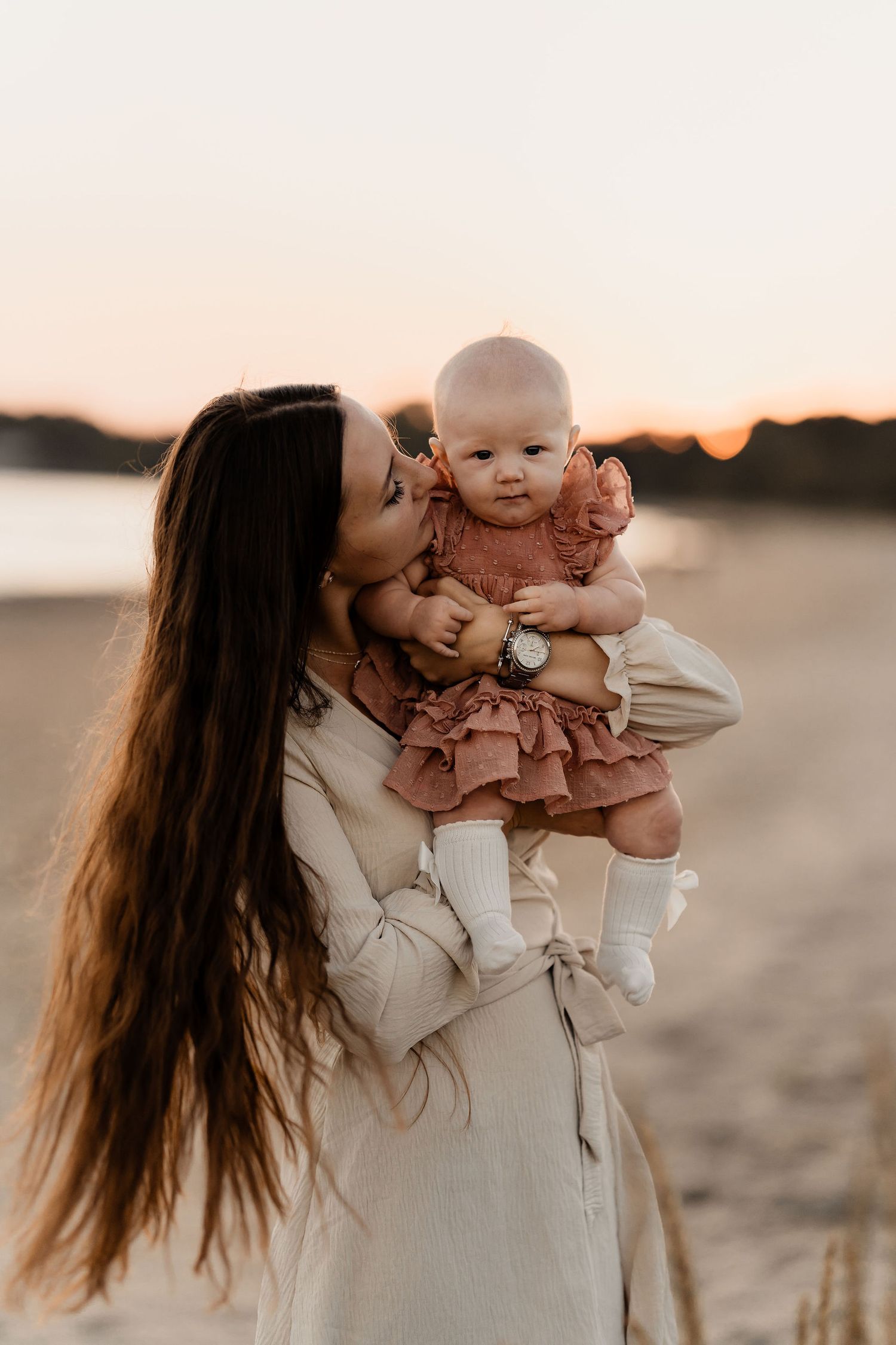 A tender sunset moment captured between parent and baby on a beach with warm golden light.