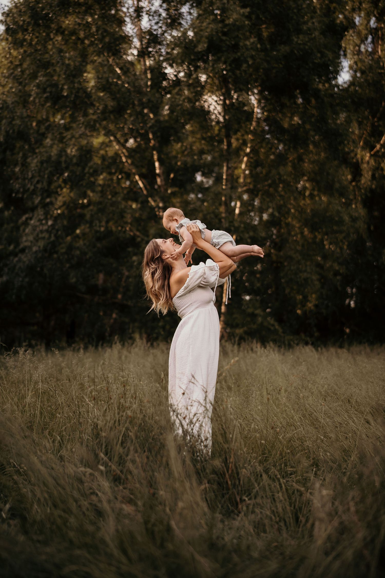 A figure in a long white dress lifts a small child playfully in a meadow at sunset against a backdrop of birch trees.