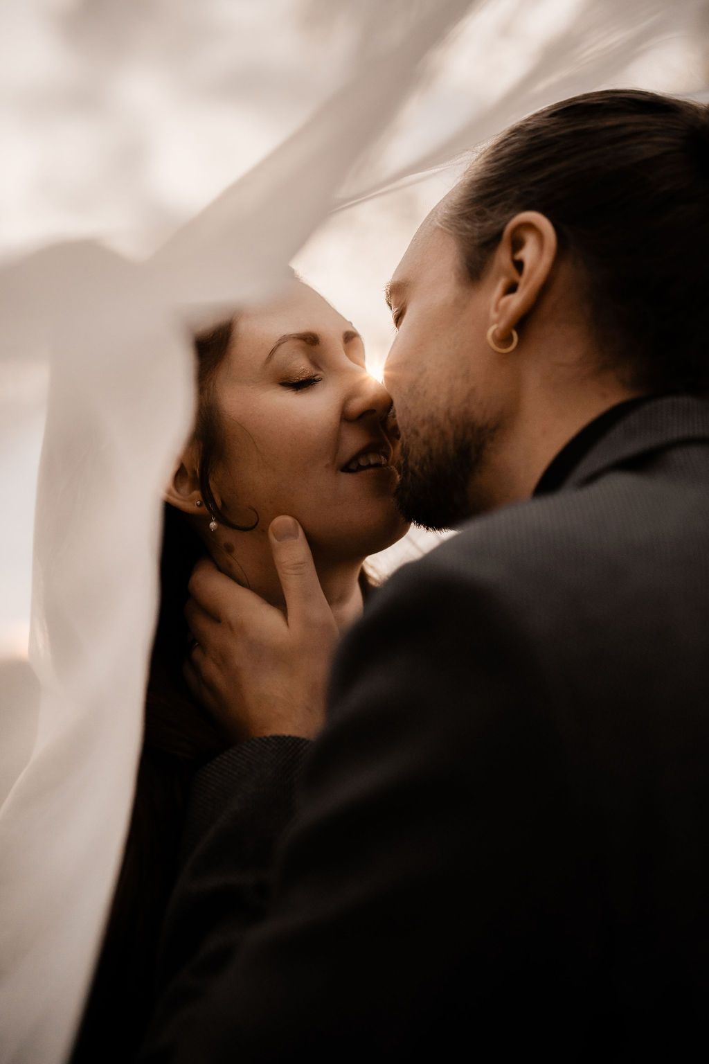 A romantic couple shares an intimate kiss under a flowing veil during their wedding photoshoot.