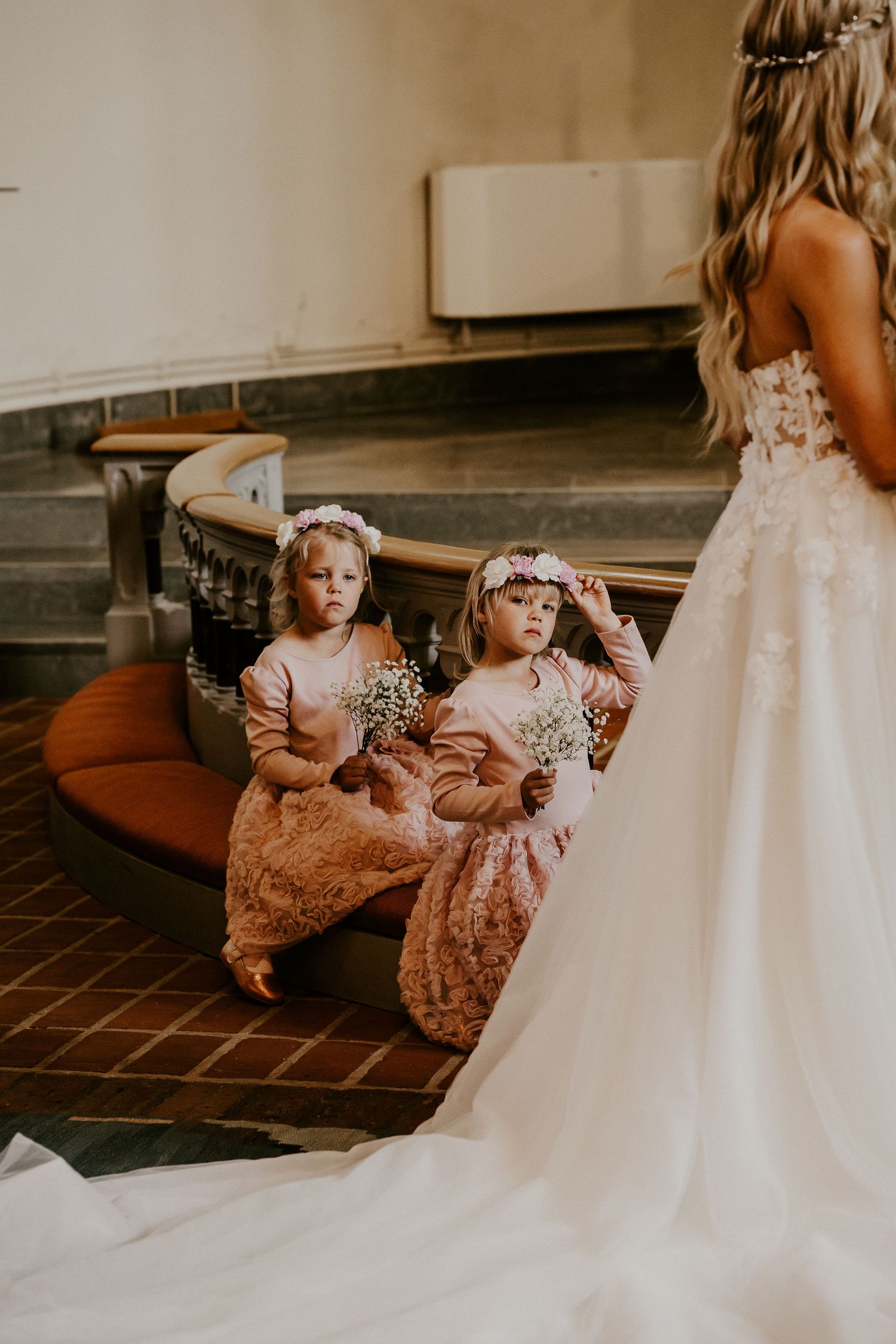 Two flower girls in rose gold dresses sit with white floral crowns as a bride's wedding gown flows beside them.