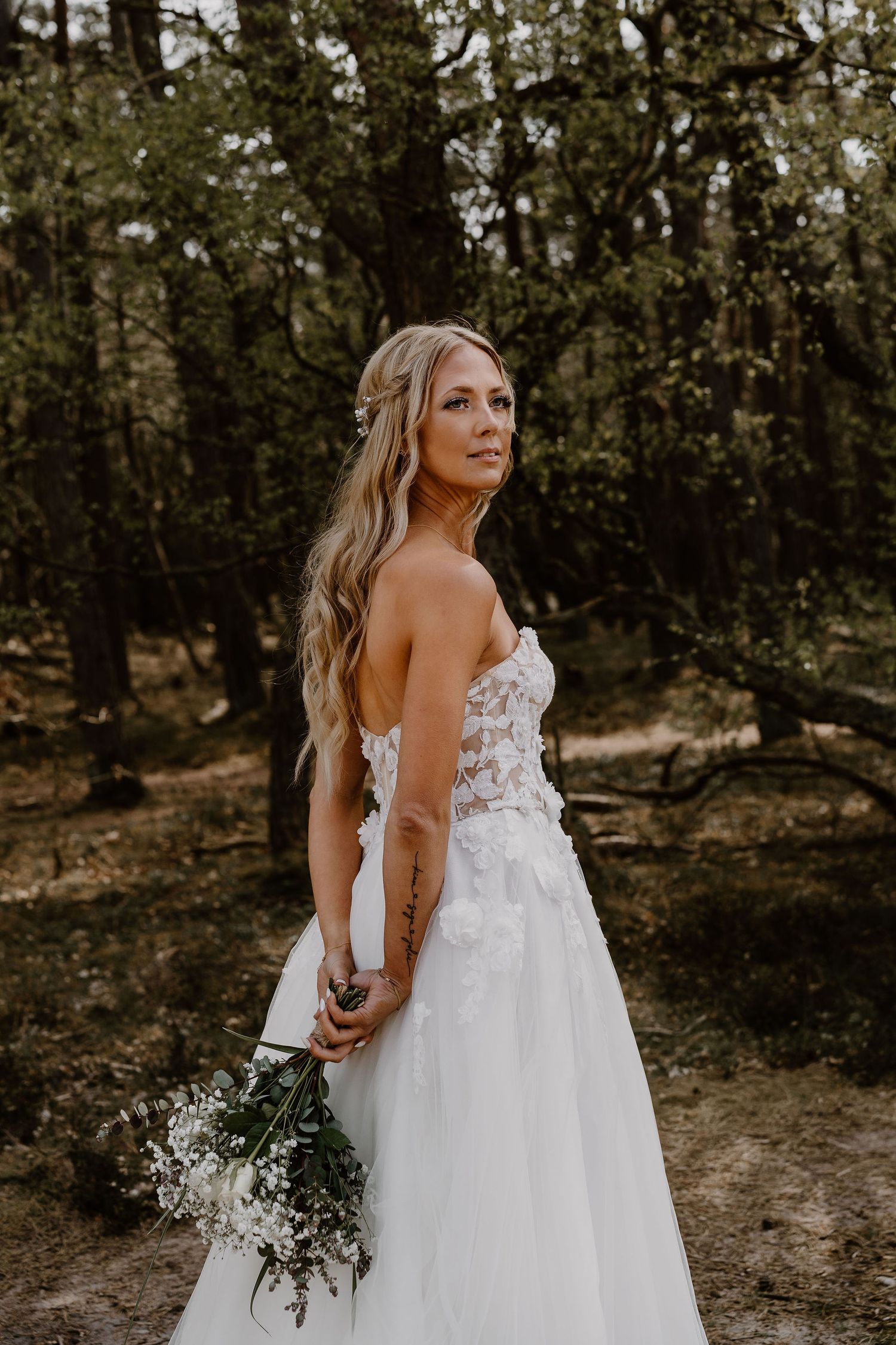 A bride in a strapless white lace wedding gown poses in a forest setting while holding a natural bouquet.