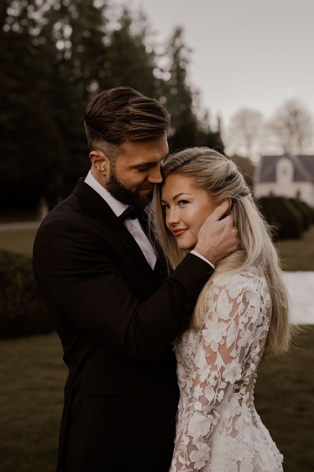 A couple shares an intimate moment in their wedding attire on a moody outdoor lawn with trees in the background.