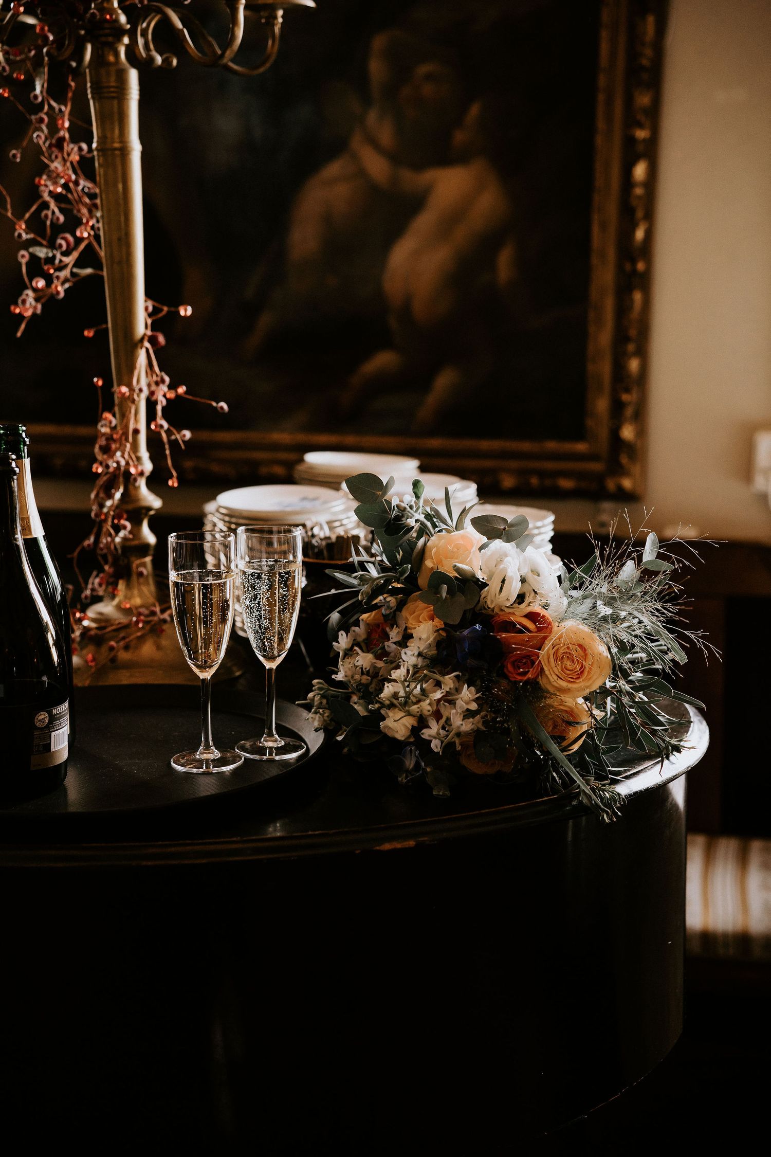 Two champagne flutes and a romantic floral arrangement with roses sit atop a dark table in moody lighting.