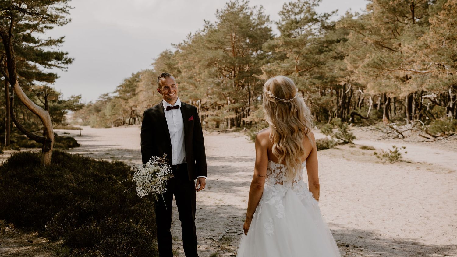 A couple stands together on a wooded path, the dress flowing in the gentle breeze among pine trees.