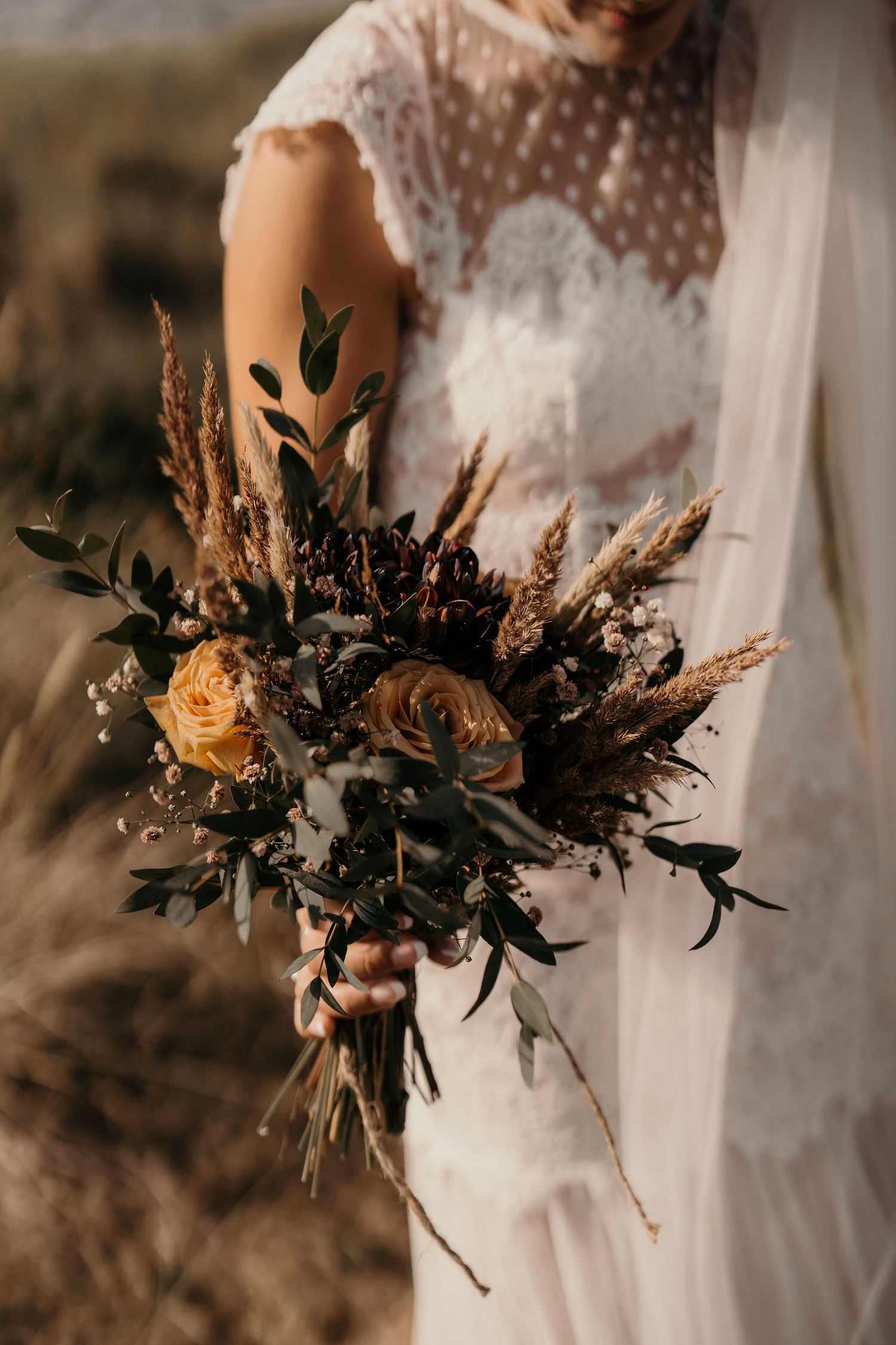 A rustic wedding bouquet with yellow roses, dried pampas grass, and dark greenery against a white lace dress.