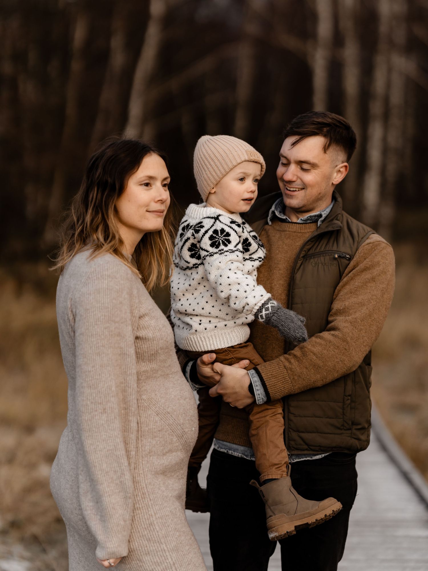 Family of three poses together on wooden boardwalk during autumn season.