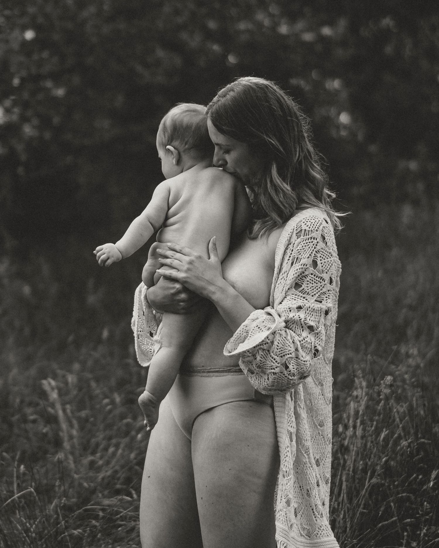 Intimate portrait of a parent in lace cardigan holding a baby outdoors.