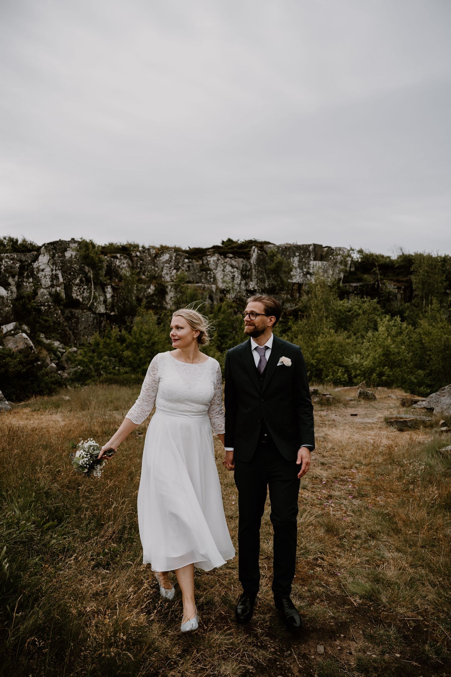 A couple in wedding attire walking together across a scenic rocky terrain on an overcast day.