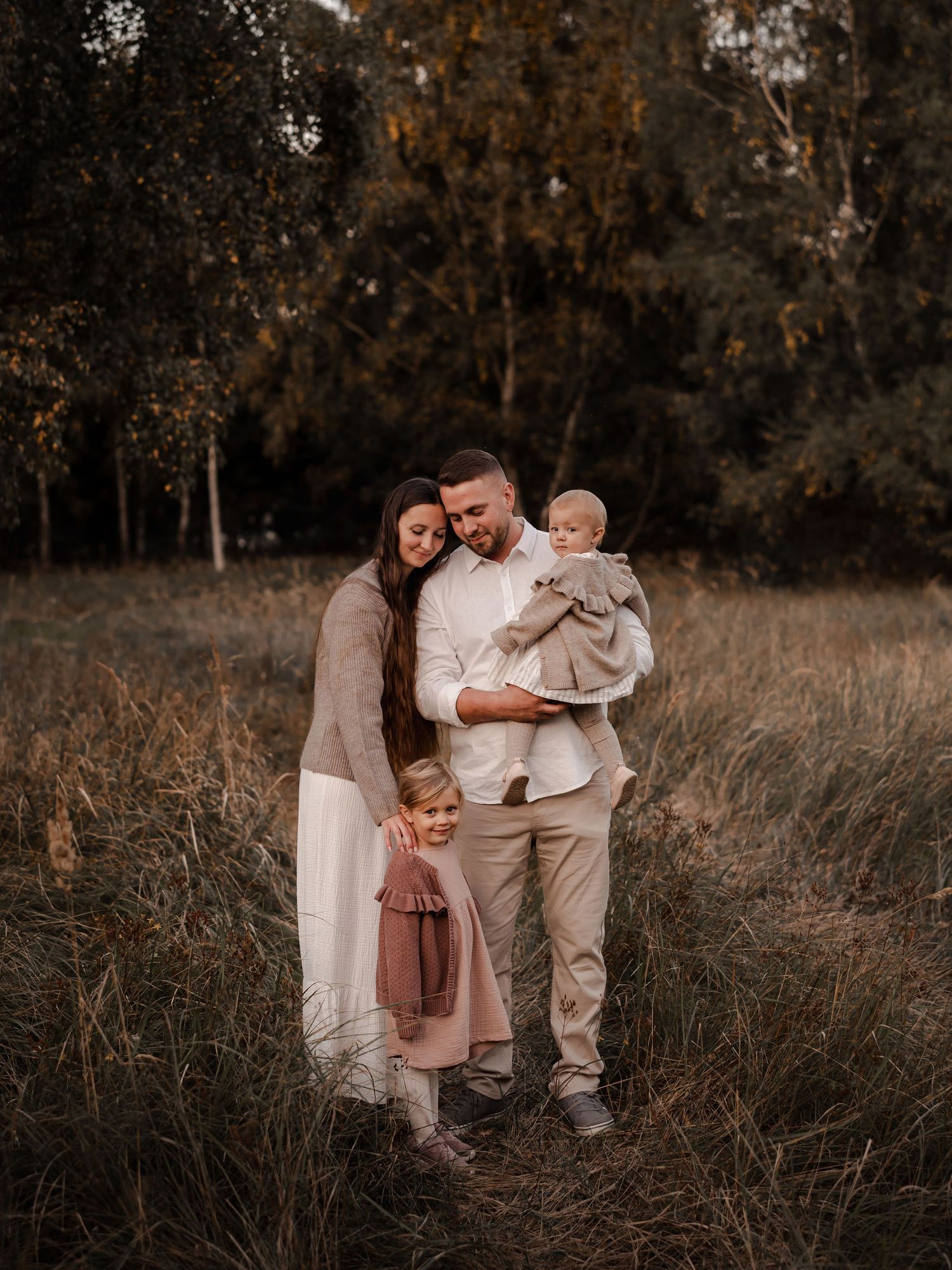 Family of three poses together in autumn field wearing coordinated neutral tones.